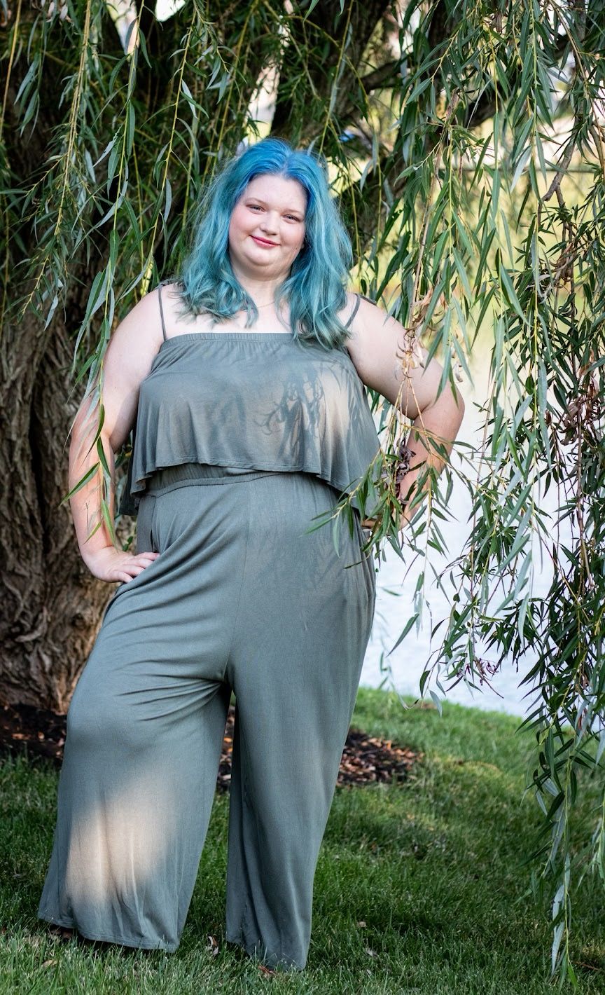 A person with blue-green hair poses under willow tree branches wearing a grey off-shoulder dress. Senior session Cincinnati