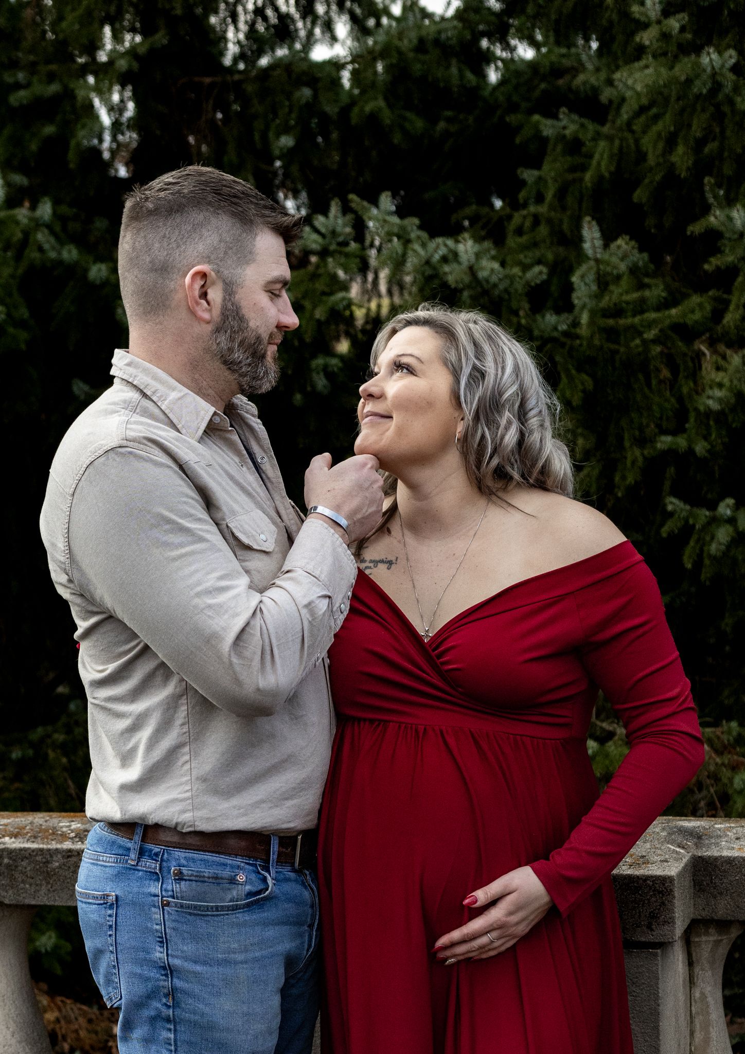 Expectant parents share an intimate moment outdoors, with the woman wearing a red dress and the man in casual attire.