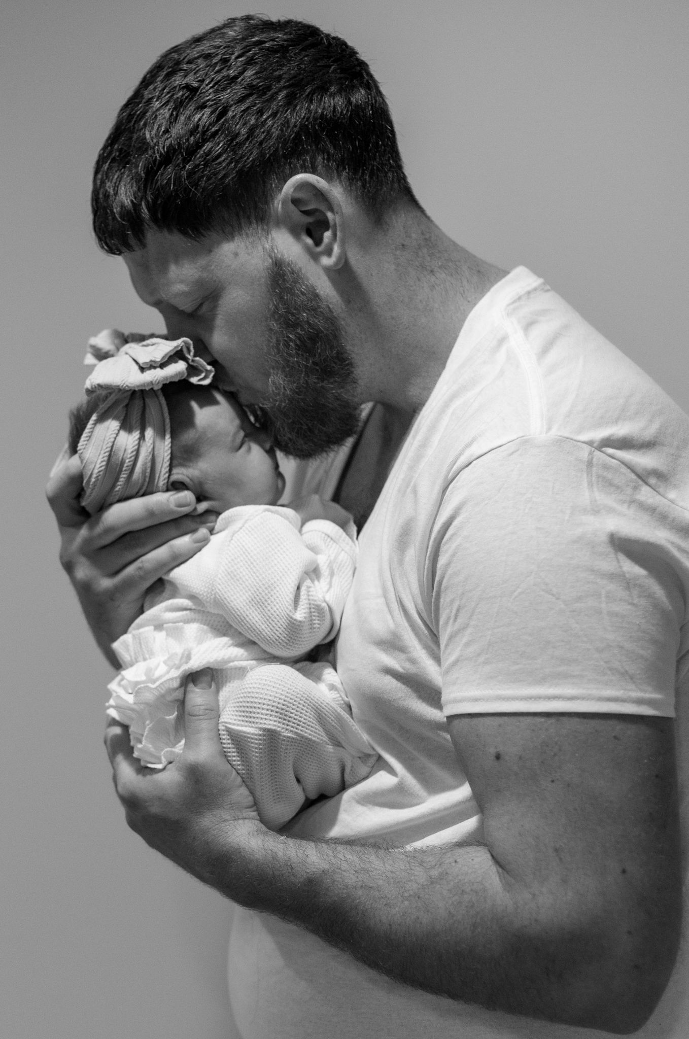 A tender black and white portrait of a fatherin a white t-shirt holding and kissing a newborn baby.
