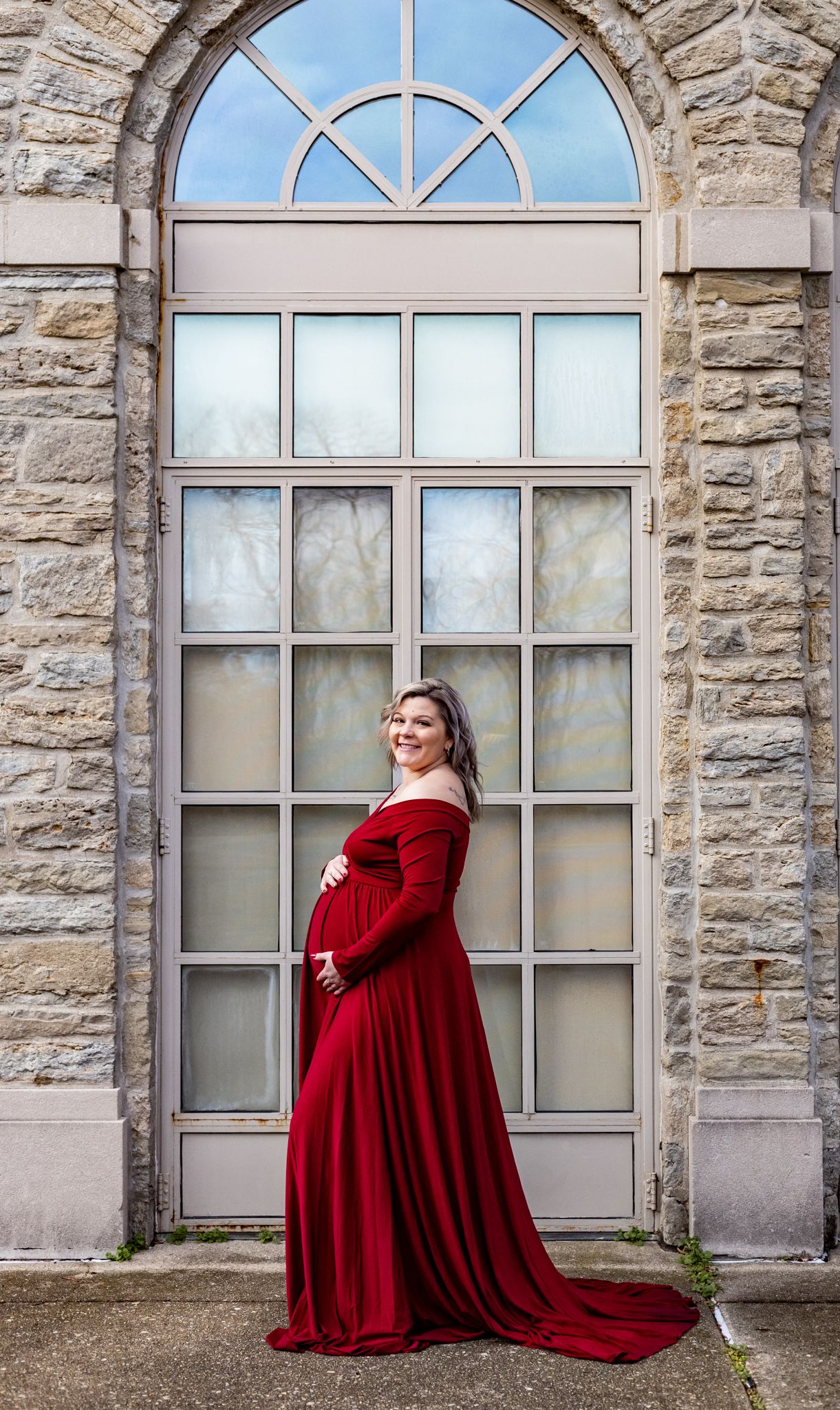 Photo of a pregnant woman in a red velvet dress posing dramatically against a stone building with arched windows.