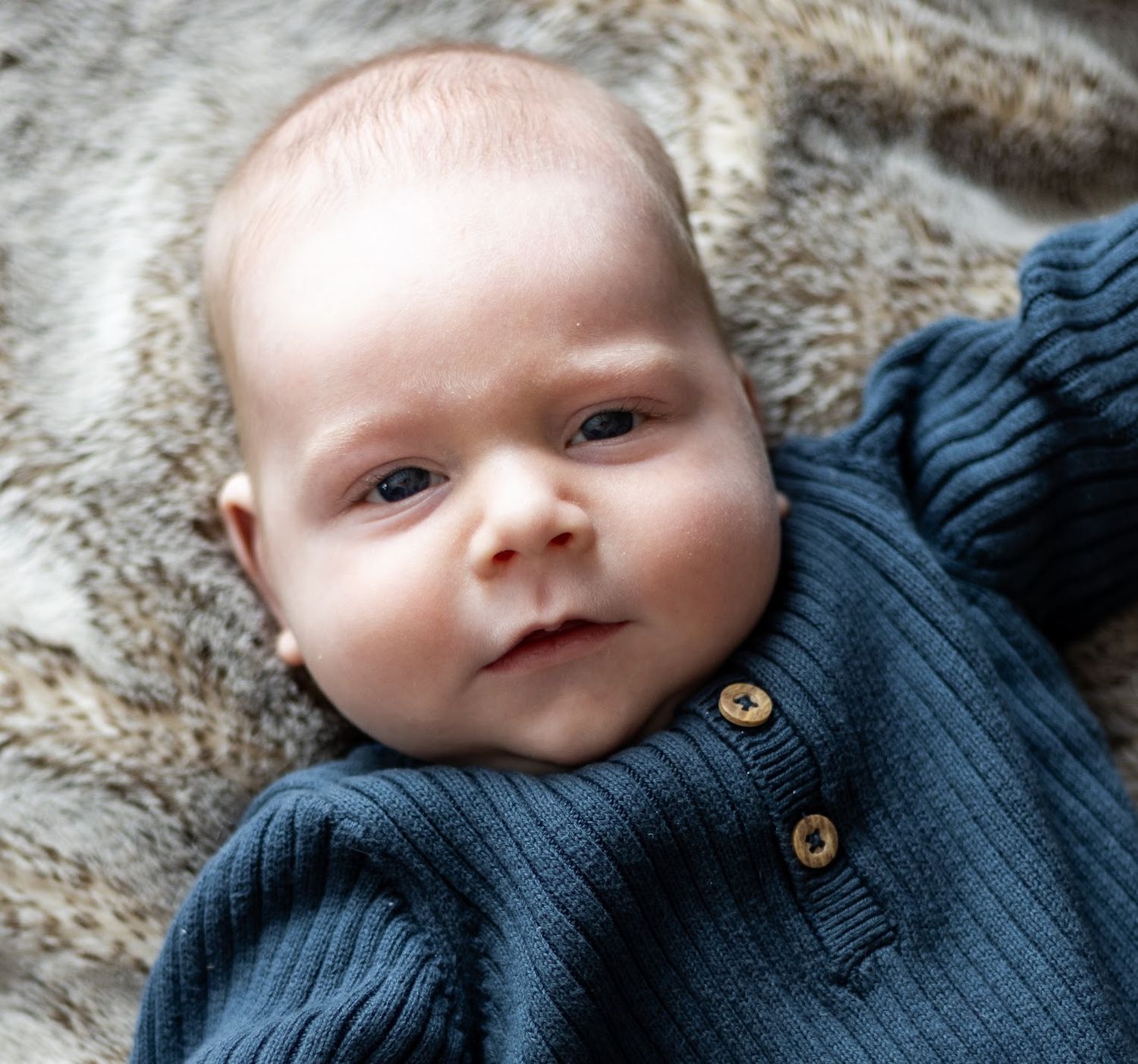 A baby wearing a navy blue cardigan sweater lies on a plush fur blanket. Cincinnati/ northern Kentucky