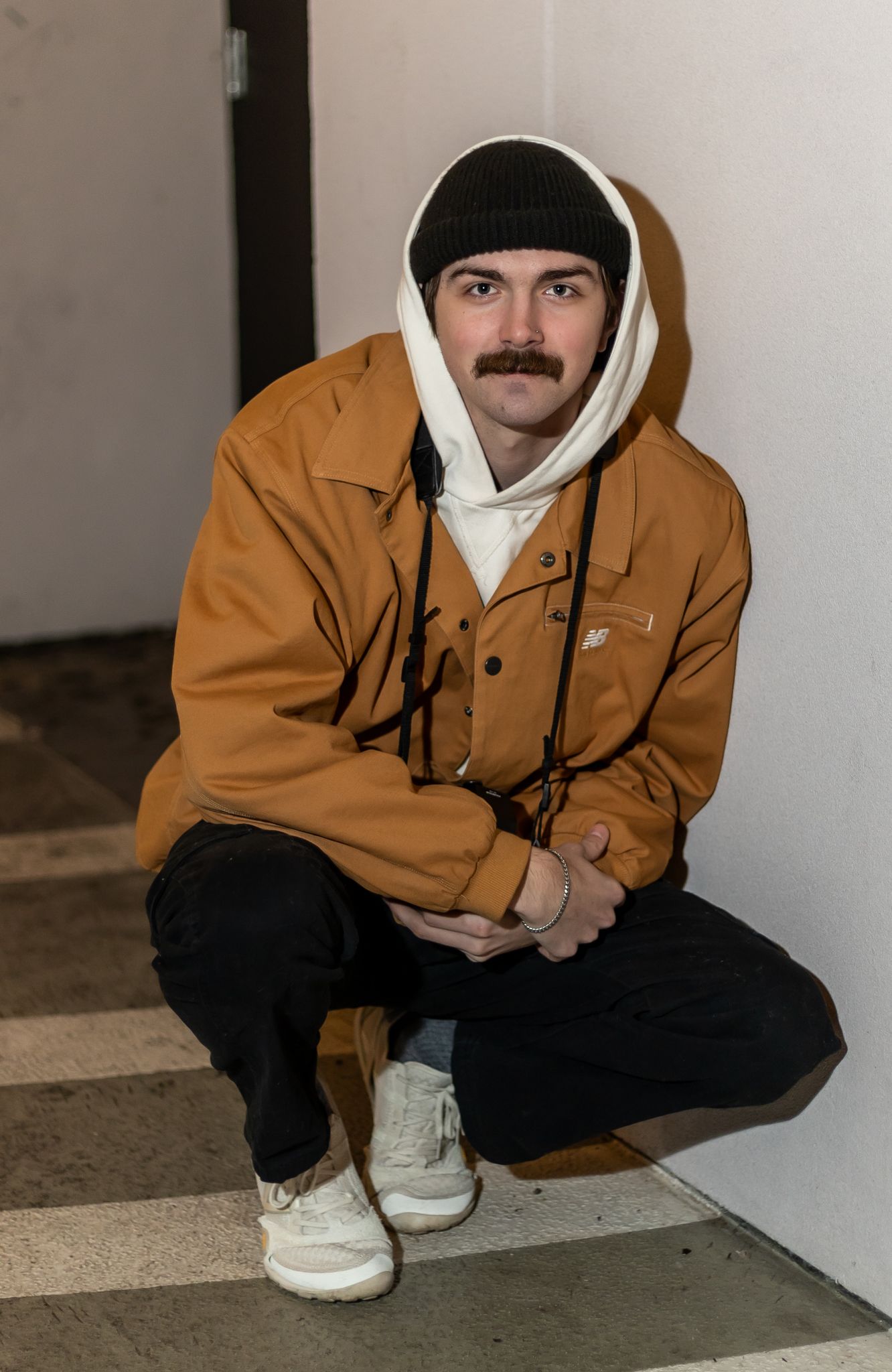 Person wearing brown jacket and black beanie crouches down showing white sneakers in minimalist setting. Cincinnati/ northern Kentucky