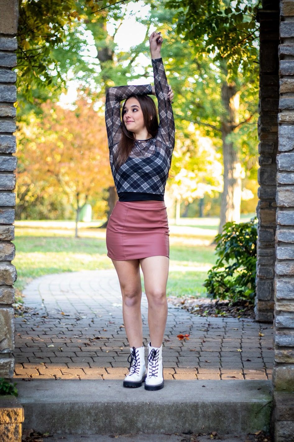 A person in a plaid crop top and pink skirt poses between stone pillars on a brick path during autumn.