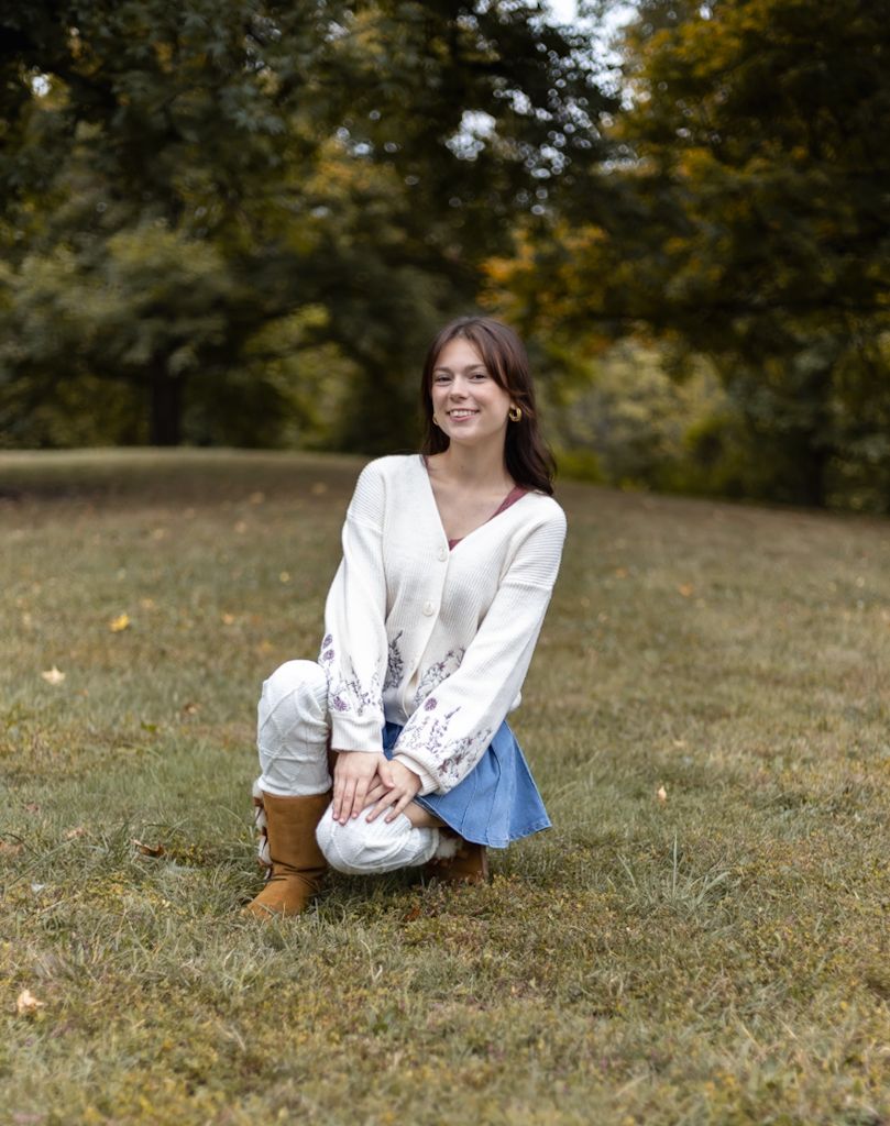 A person in a white blouse sits on grass in an autumn outdoor setting during a casual photoshoot. Cincinnati/ northern Kentucky
