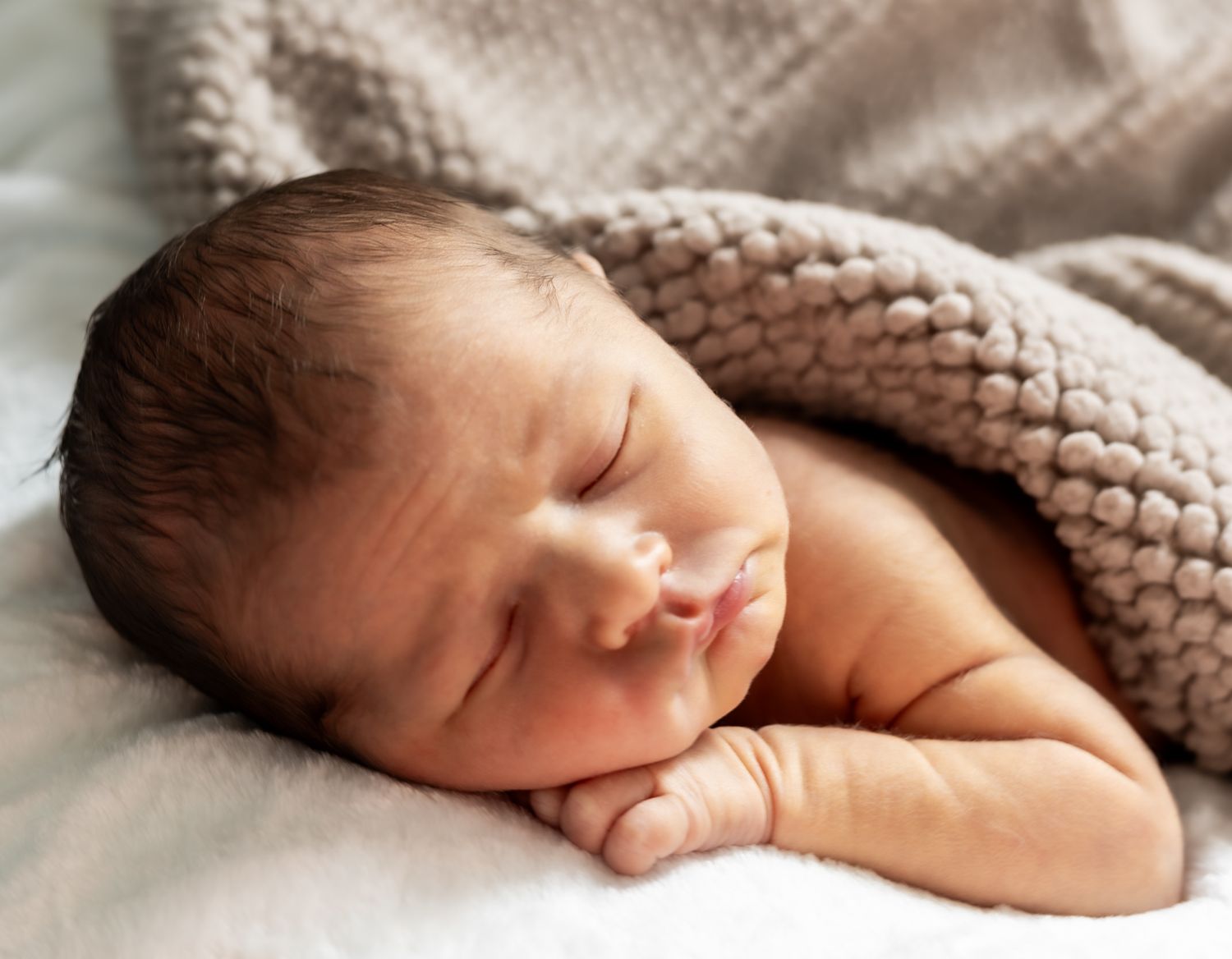 A sleeping newborn baby wrapped in a soft knitted blanket rests peacefully on white bedding. Cincinnati/ Northern Kentucky