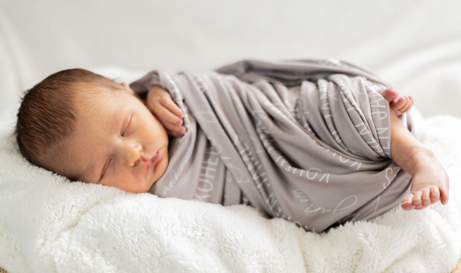 A sleeping baby wearing grey clothing rests peacefully on white bedding. Cincinnati/ Northern Kentucky