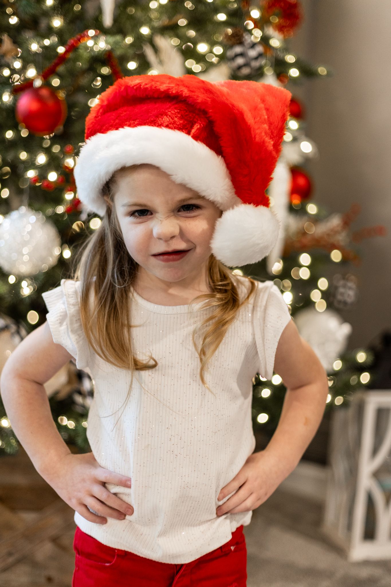 A child in a white sweater and Santa hat poses playfully in front of a decorated Christmas tree. Cincinnati/ northern Kentucky