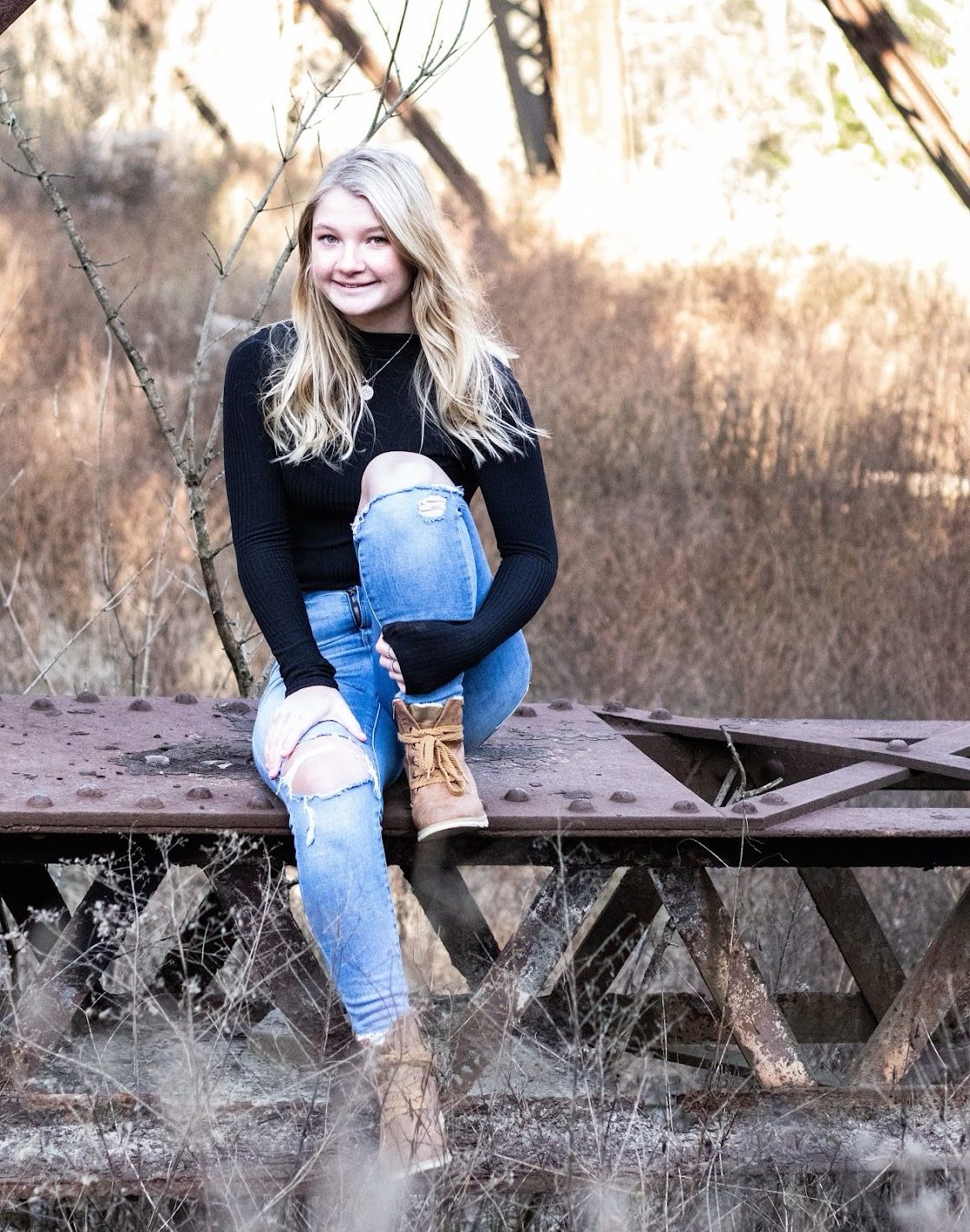 Person in jeans and black sweater sitting casually on rustic wooden bench surrounded by dried winter grasses.