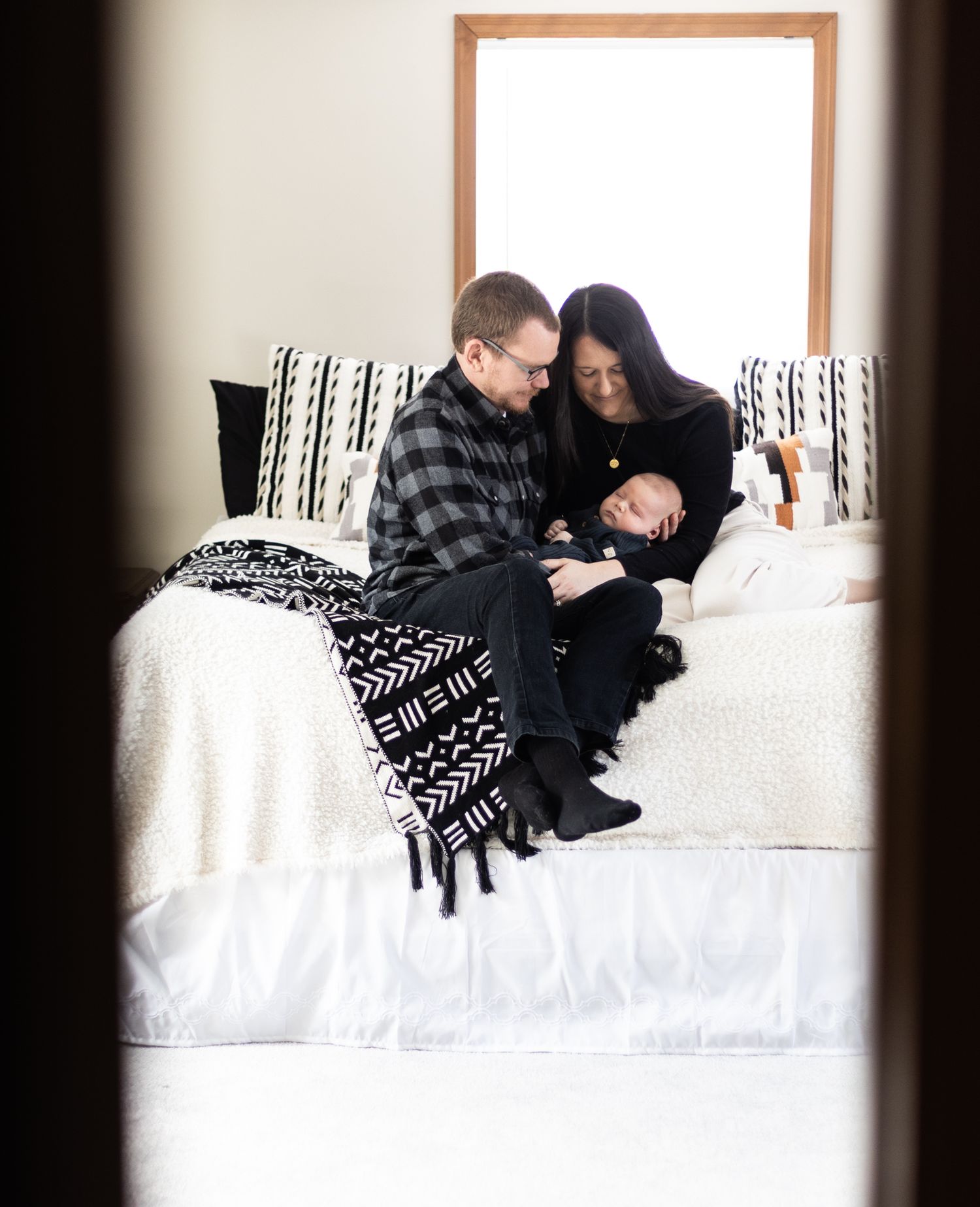 A couple in cozy winter sweaters cuddles together on a white bed in a softly lit room. Cincinnati/ Northern Kentucky