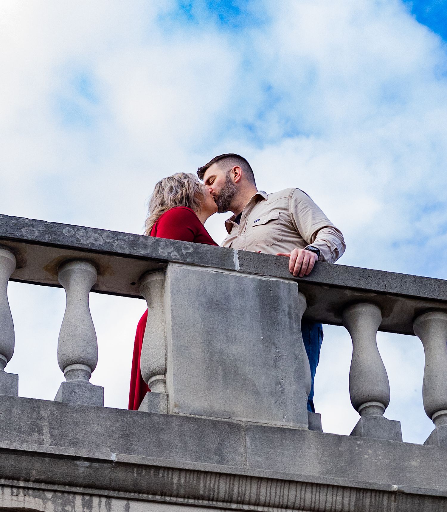A couple shares a romantic moment on a stone balcony against a blue cloudy sky. Cincinnati/ Northern Kentucky