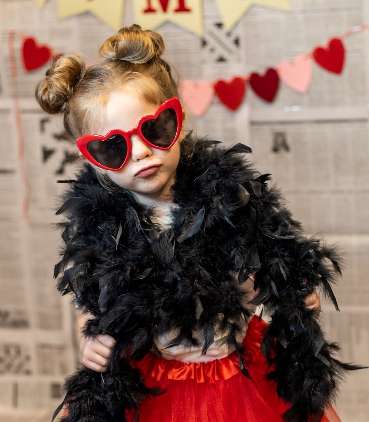 A stylish young model wearing heart-shaped red sunglasses, black feather boa and red dress poses against a Valentine's backdrop.