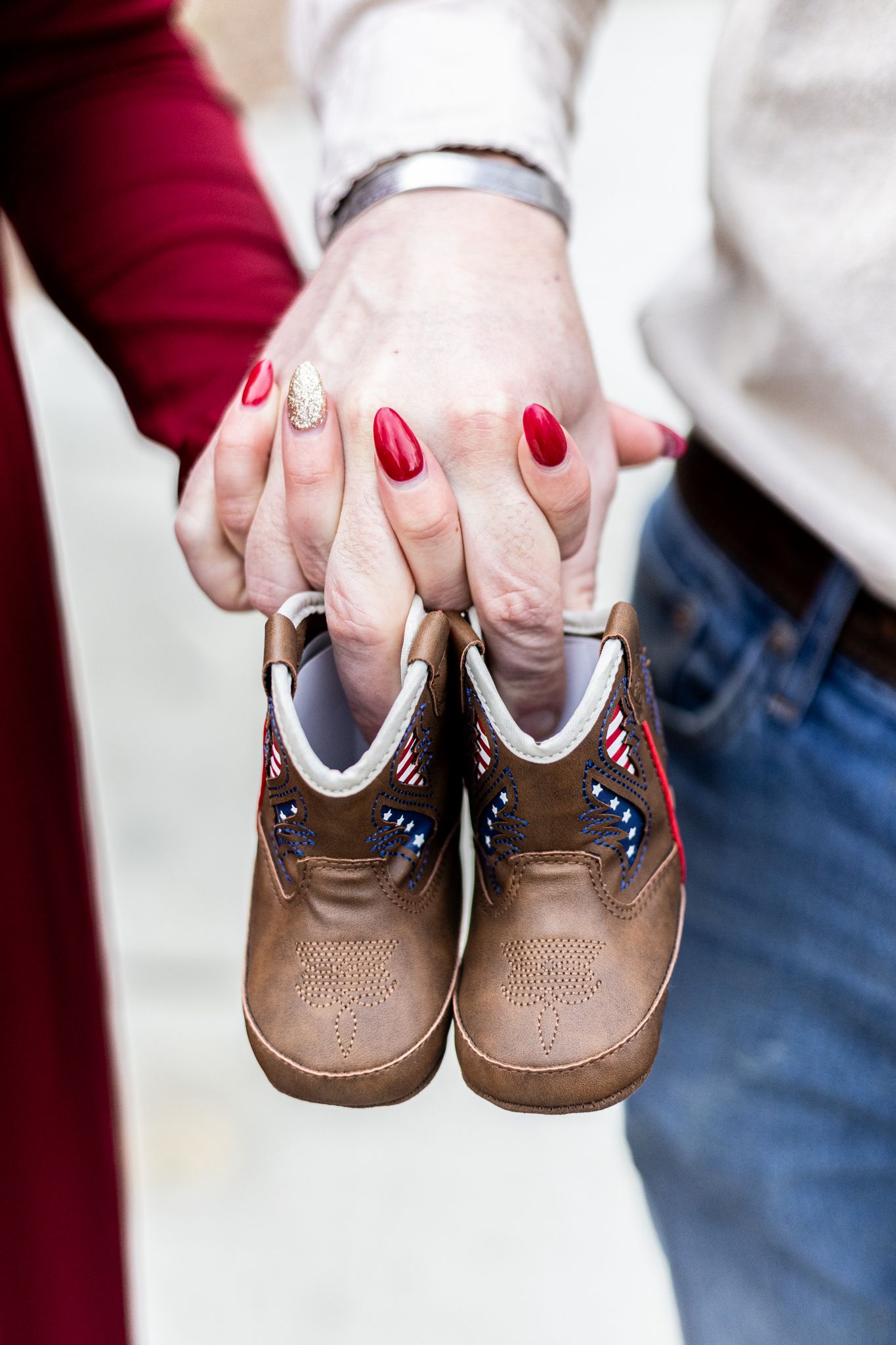 Hands with red nail polish hold small brown leather baby shoes against a white background. Cincinnati/ Northern Kentucky