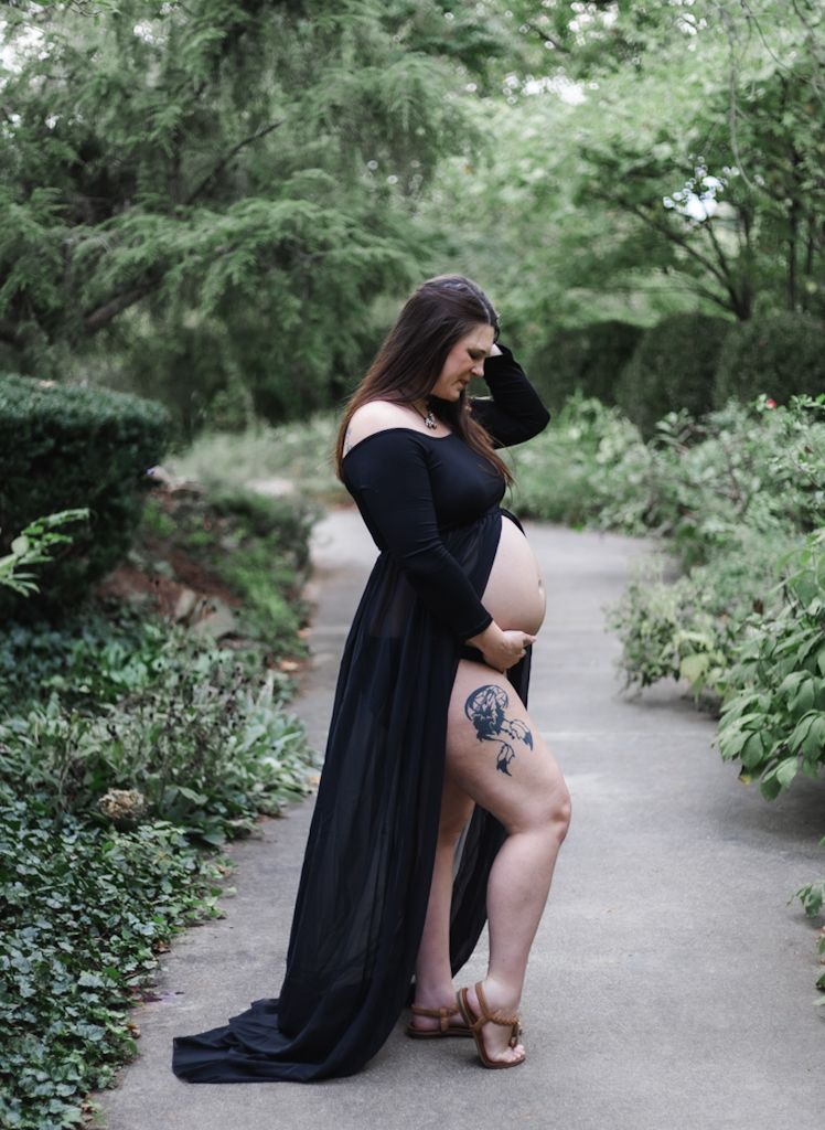 Pregnant woman in black off-shoulder gown with thigh slit and floral tattoo posing in lush garden pathway.
