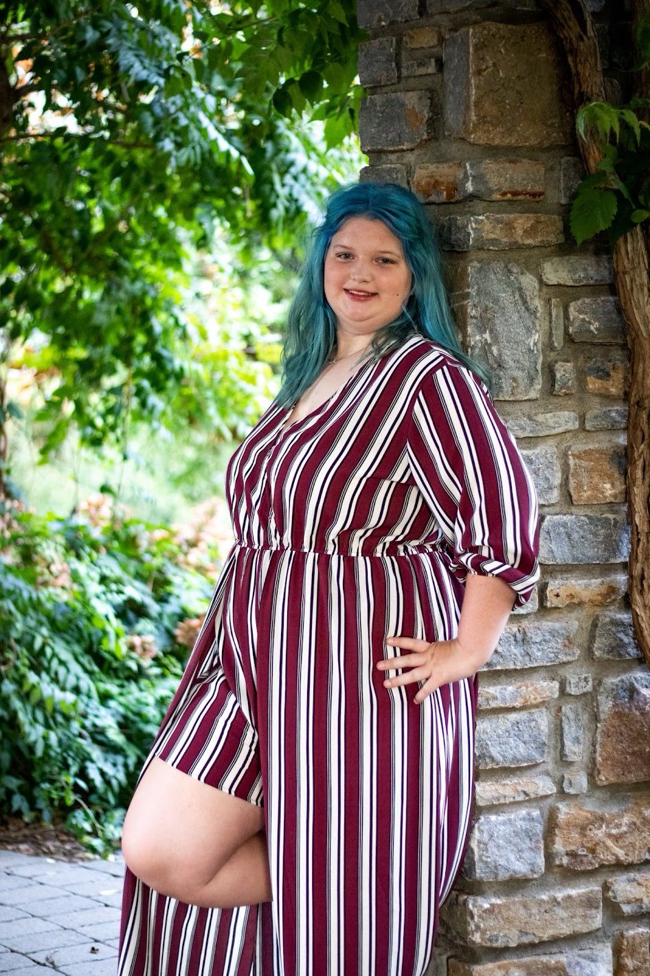 A person in a burgundy striped dress poses in front of a stone wall with green foliage in the background. Female senior Cincinnati/