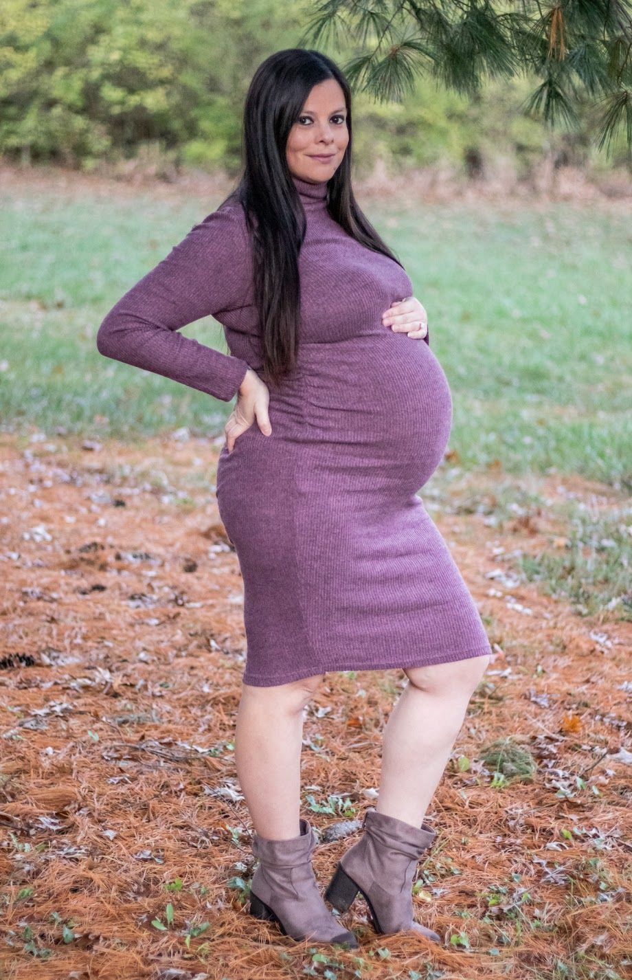 A maternity photo of a woman in a purple sweater dress and brown ankle boots poses in autumn leaves on the ground.