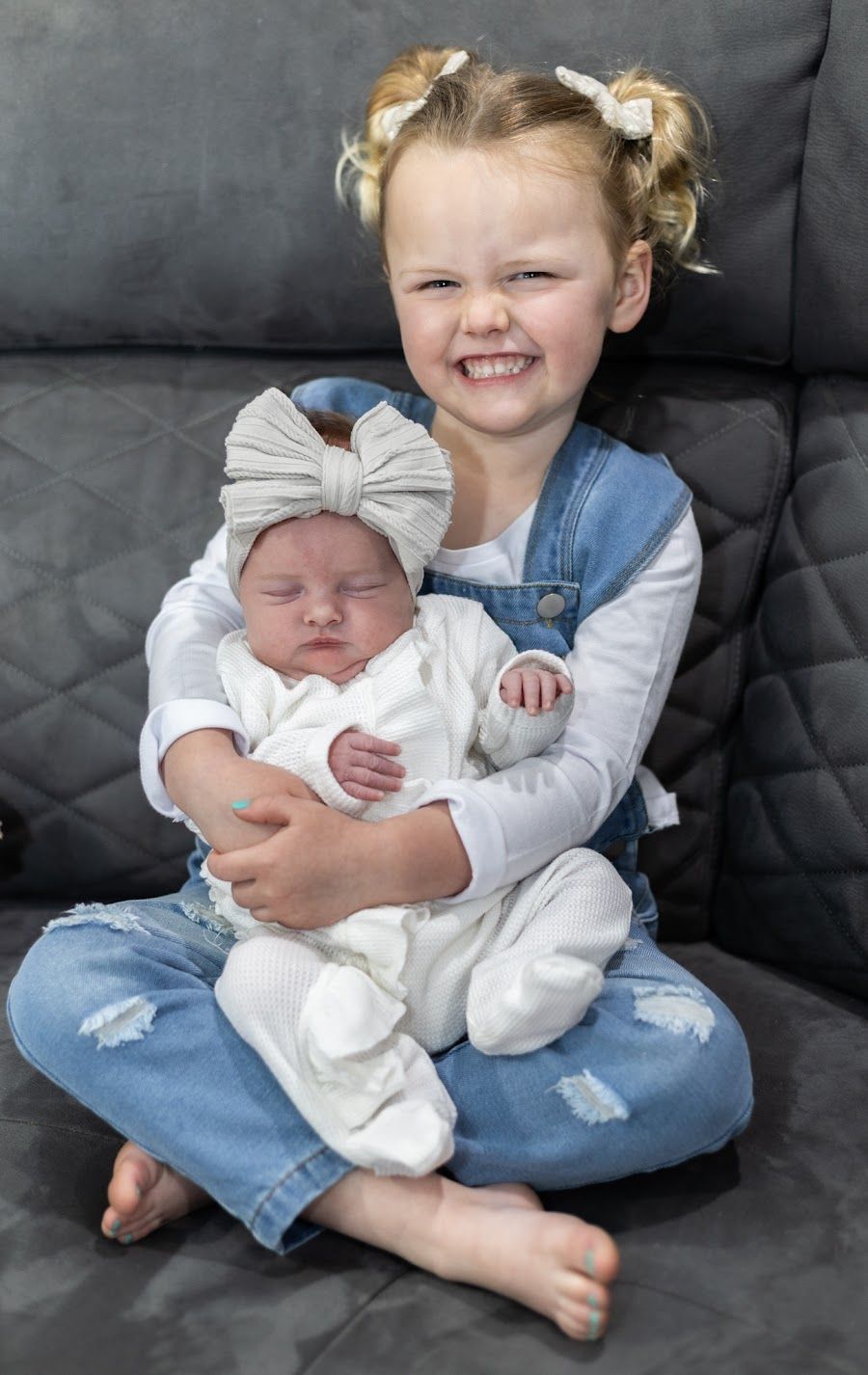 A photo showing a young child in ripped jeans holding a newborn baby wearing white on a dark couch.