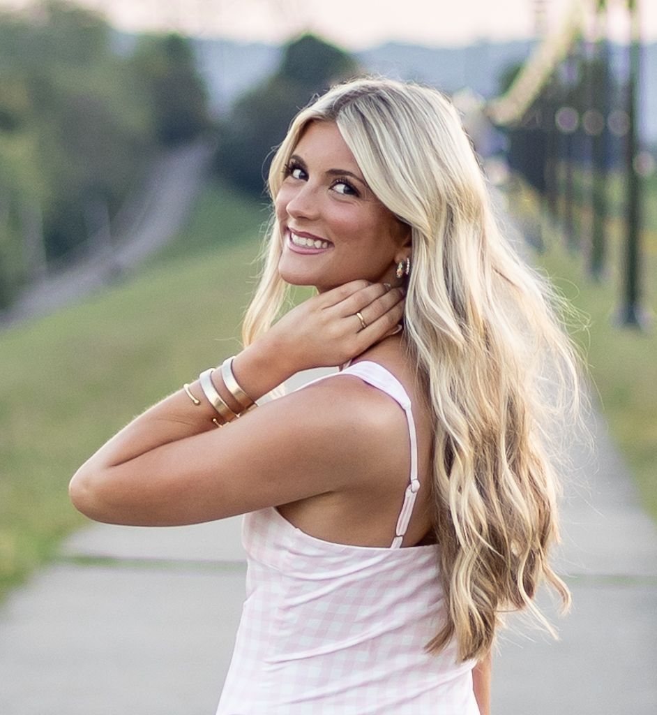 A woman with long blonde wavy hair smiles while wearing a white tank top on a path with utility poles in the background. Cincinnati