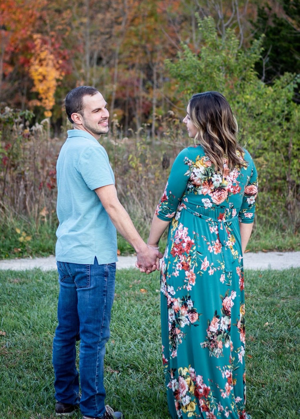 A couple in blue and teal floral dress hold hands while walking through an autumn landscape during a photo session.