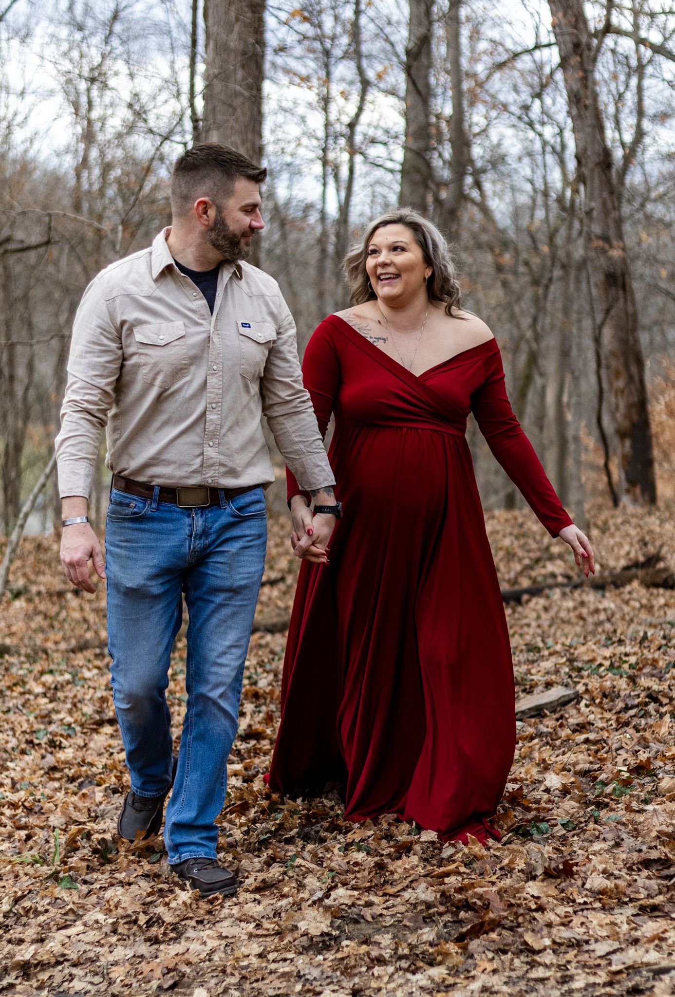 A couple walks through autumn leaves in a forest setting wearing burgundy dress and tan shirt with blue jeans.