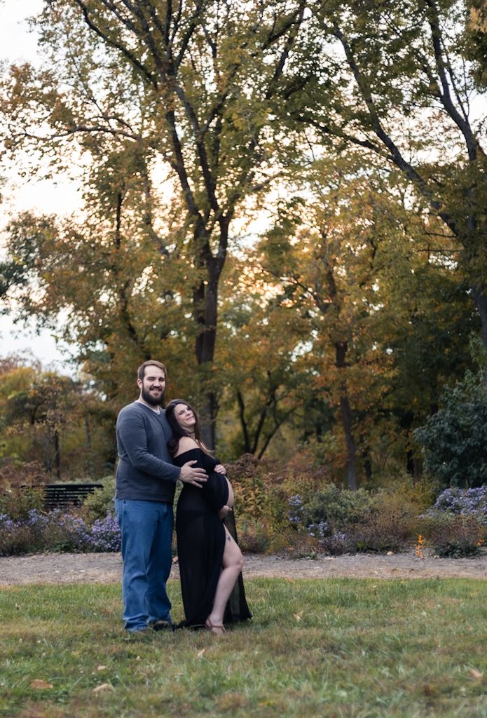Couple poses in autumn park, woman in black maternity gown cradled by partner among golden fall foliage.