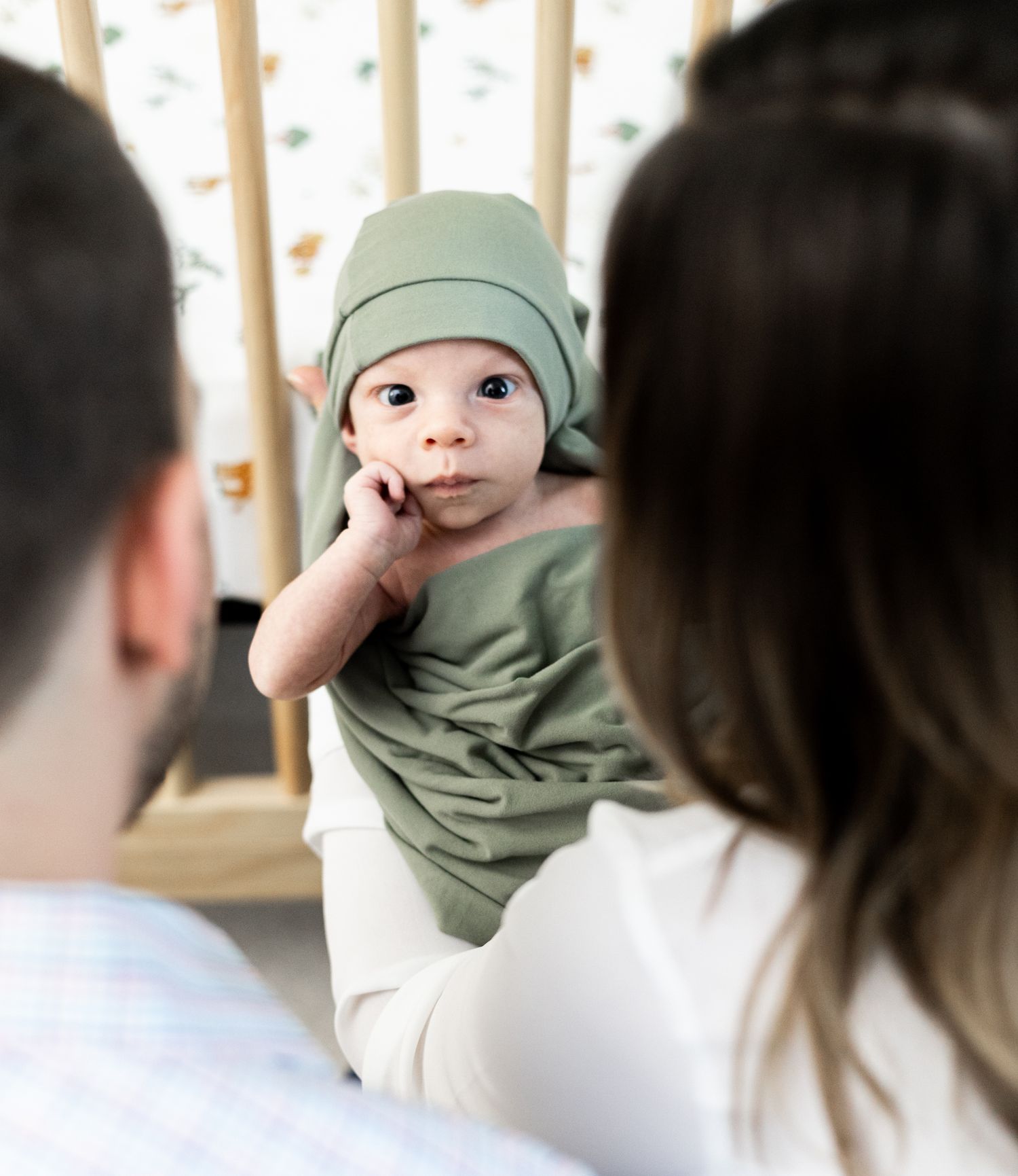 Baby wearing sage green beanie and matching shirt sits between two adults in front of a crib. Cincinnati/ Northern Kentucky