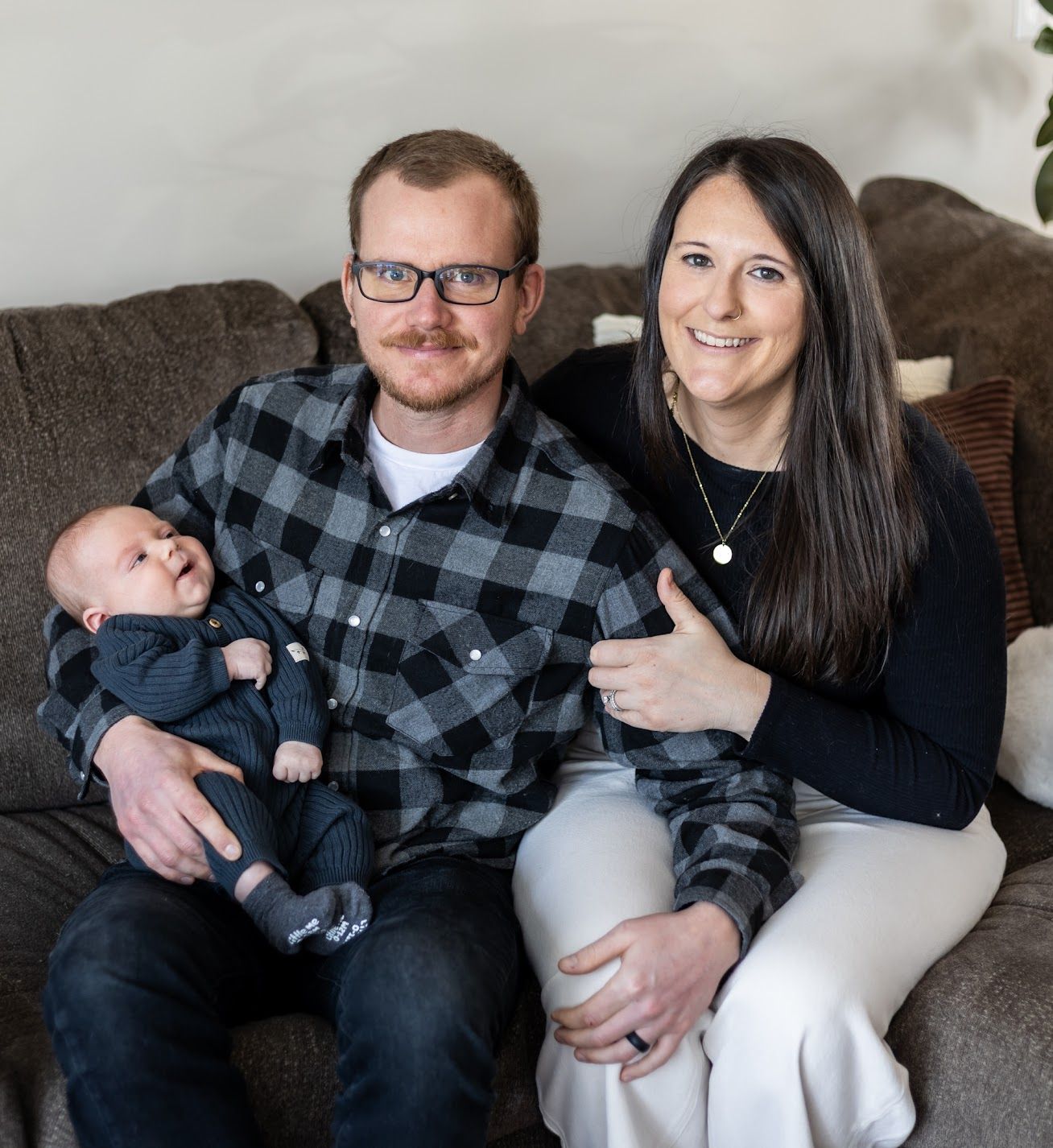 A family sits together on a gray couch, with a small baby wearing blue clothing held in a plaid flannel embrace.
