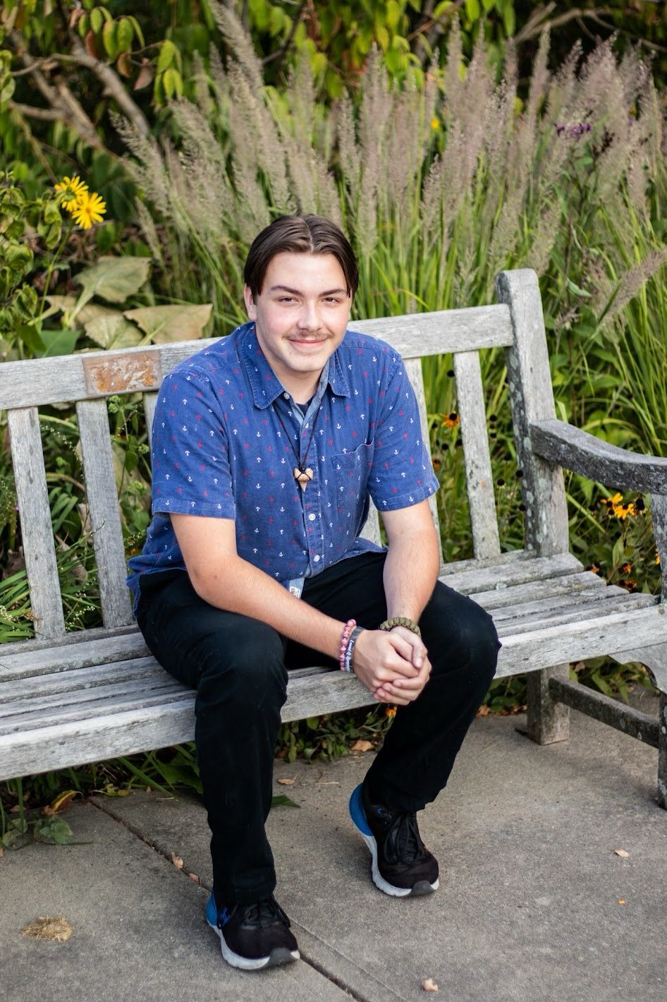 A person in a blue polka dot shirt sits on a wooden bench surrounded by ornamental grasses and garden foliage. Cincinnati/ Northern Kentucky