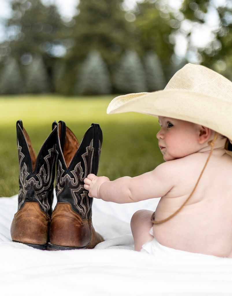 Baby in cowboy hat sitting on blanket next to brown leather cowboy boots in a field. Cincinnati/ northern Kentucky