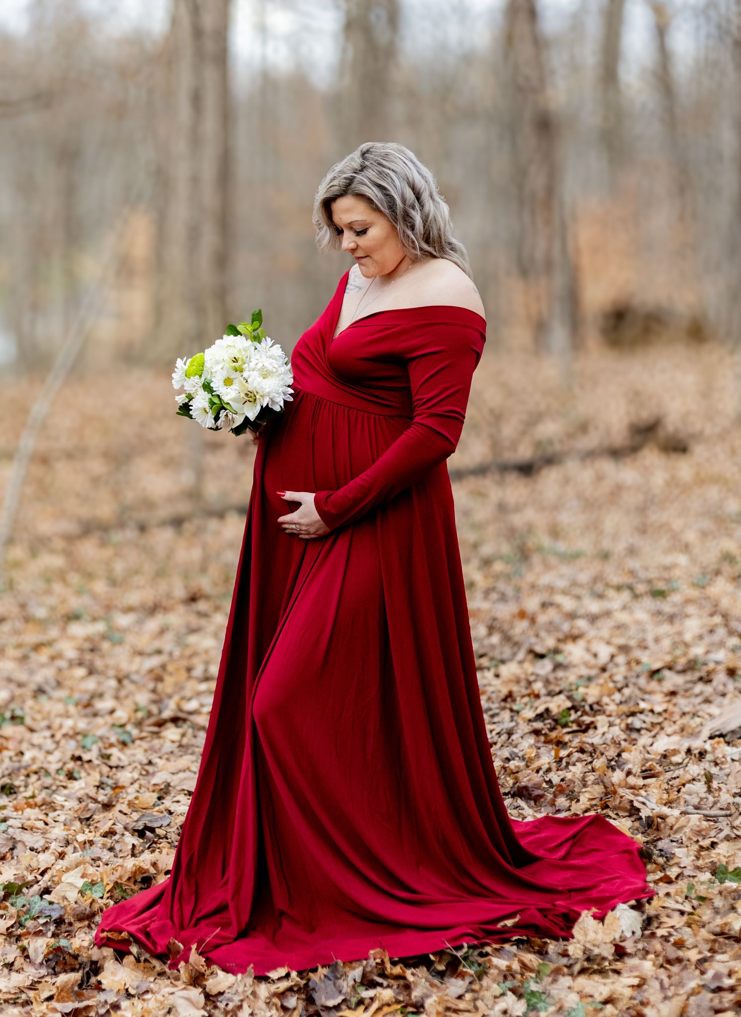 A pregnant woman in a flowing red velvet gown holds white flowers while standing on autumn leaves in a forest. Cincinnati/ Northern Kentucky