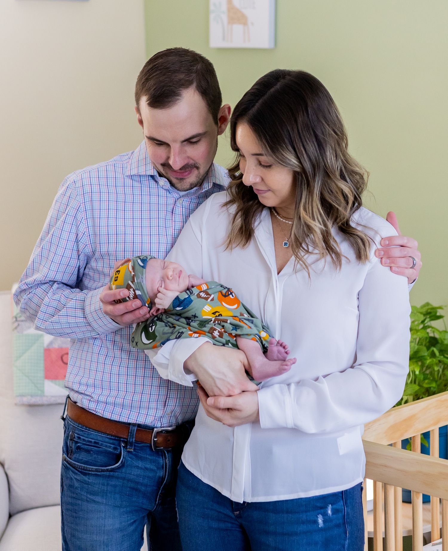A couple in a white shirt and blue plaid shirt hold and gaze lovingly at their swaddled newborn baby in a nursery setting.