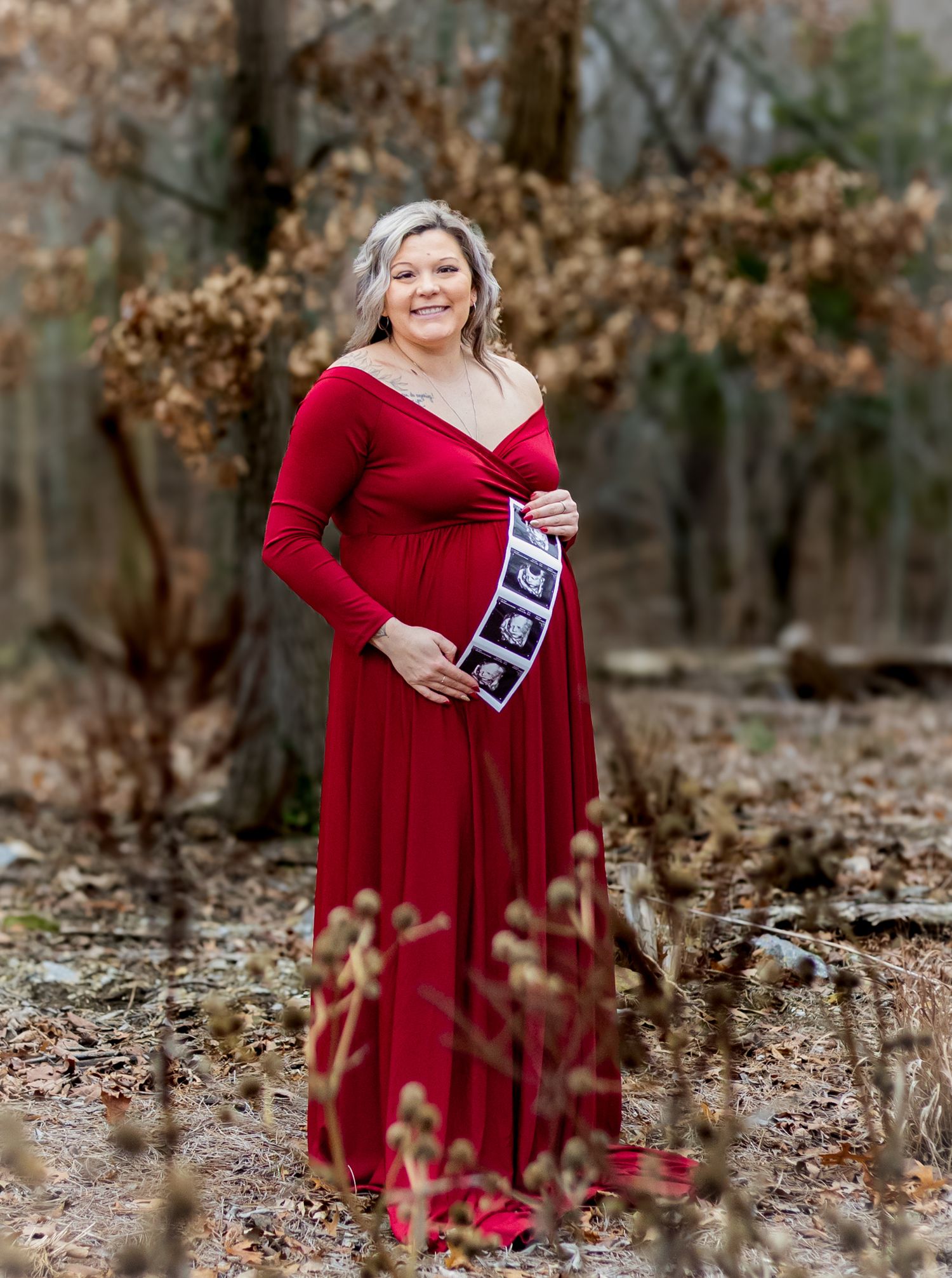 A pregnant woman in a flowing red dress poses gracefully among autumn leaves in a wooded outdoor setting. Cincinnati/ Northern Kentucky
