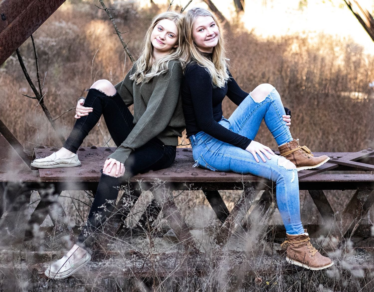 Two girls sitting on a rustic rail wearing casual clothing and sneakers in an outdoor autumn setting.