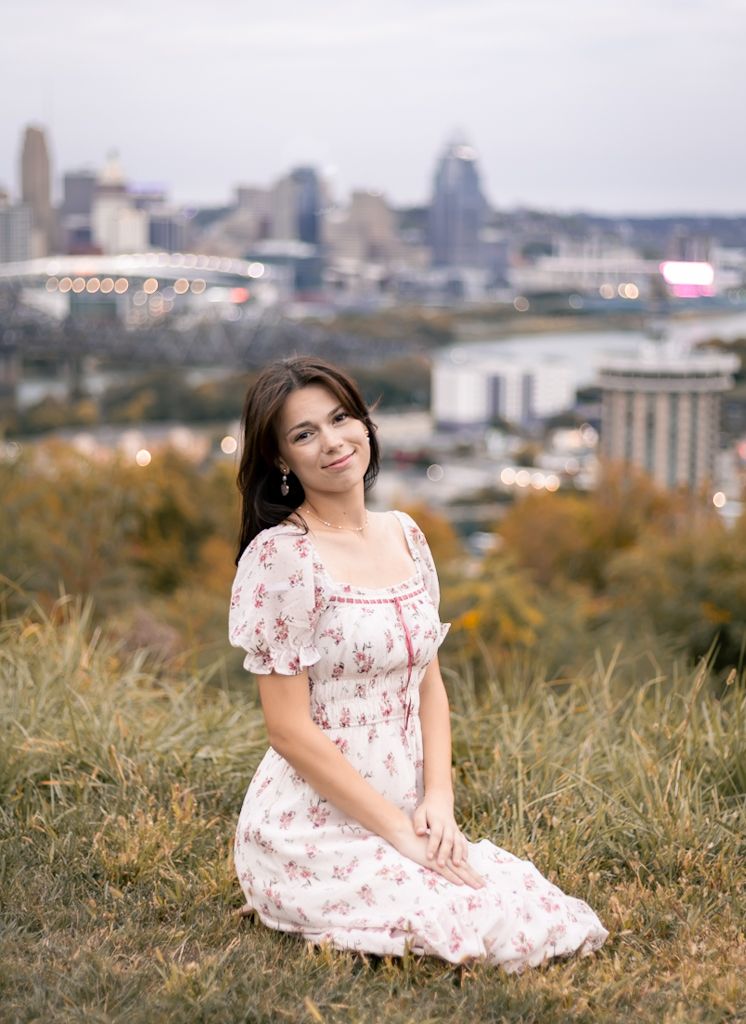 A person in a floral dress sits gracefully on a grassy hilltop with a city skyline visible in the background. Cincinnati/ northern Kentucky