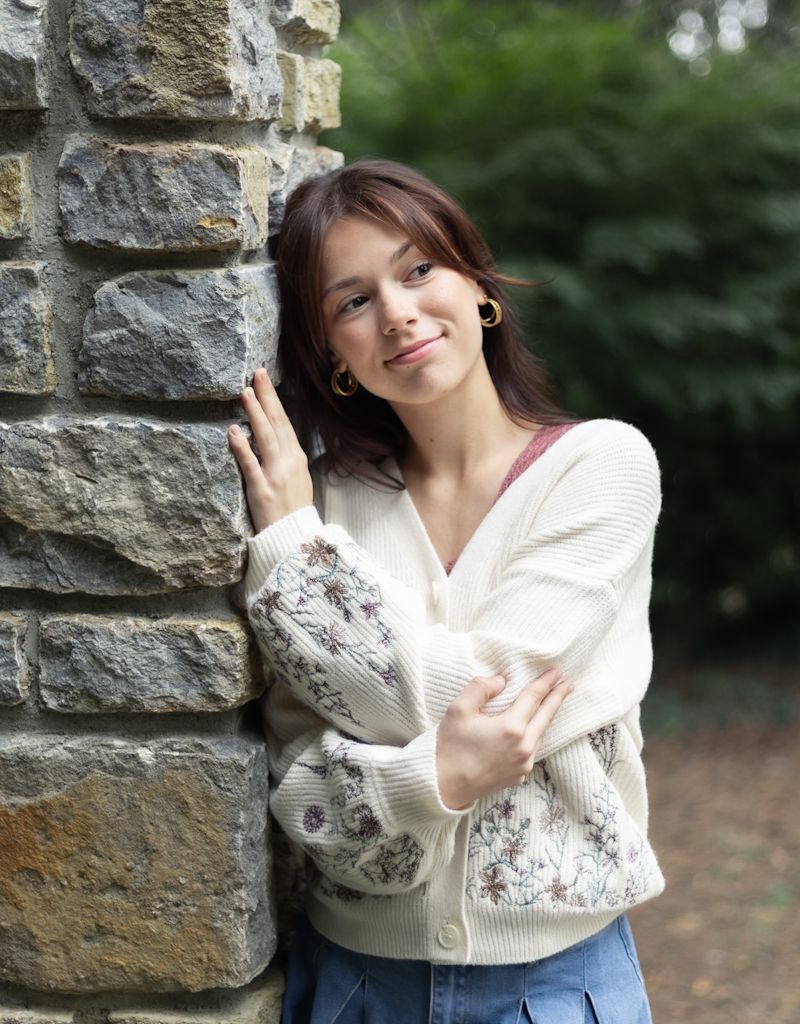 A person in a cream knit sweater poses against a stone wall in an outdoor setting. Cincinnati/ northern Kentucky