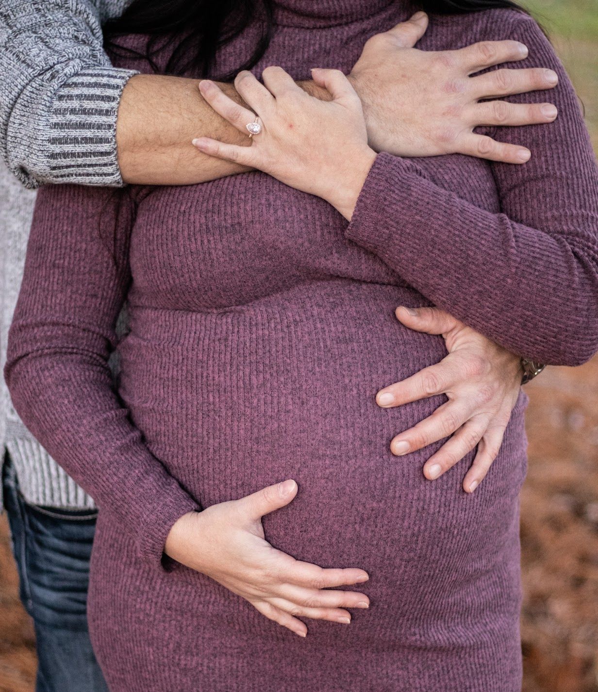 Close up of hands cradling a pregnant belly wearing a purple sweater against an autumn background.