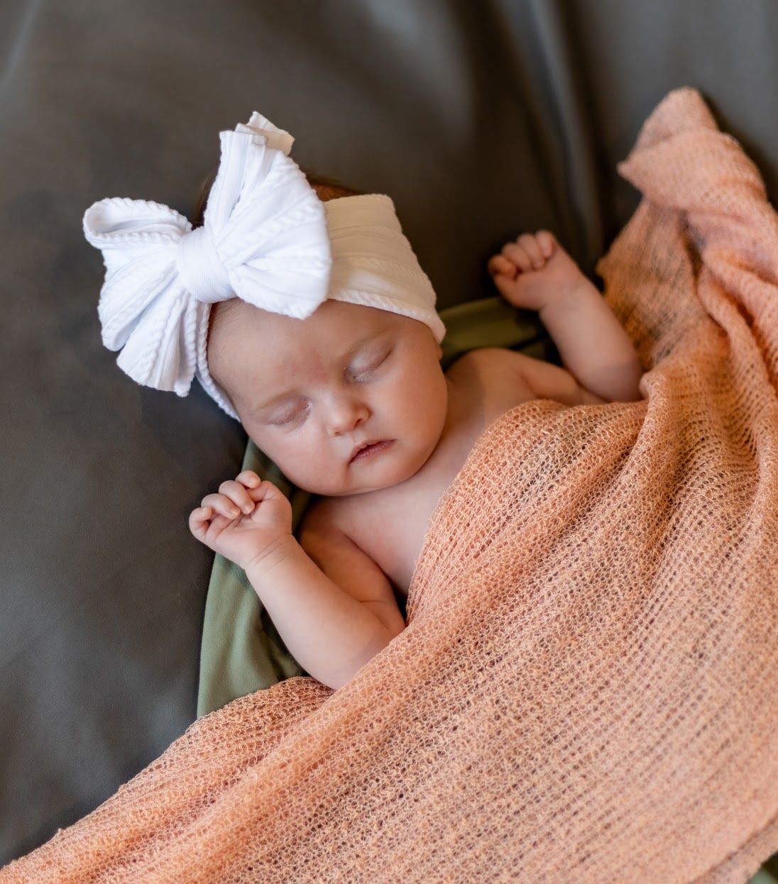 Sleeping newborn wrapped in peach-colored blanket wearing white bow headband on dark gray surface.