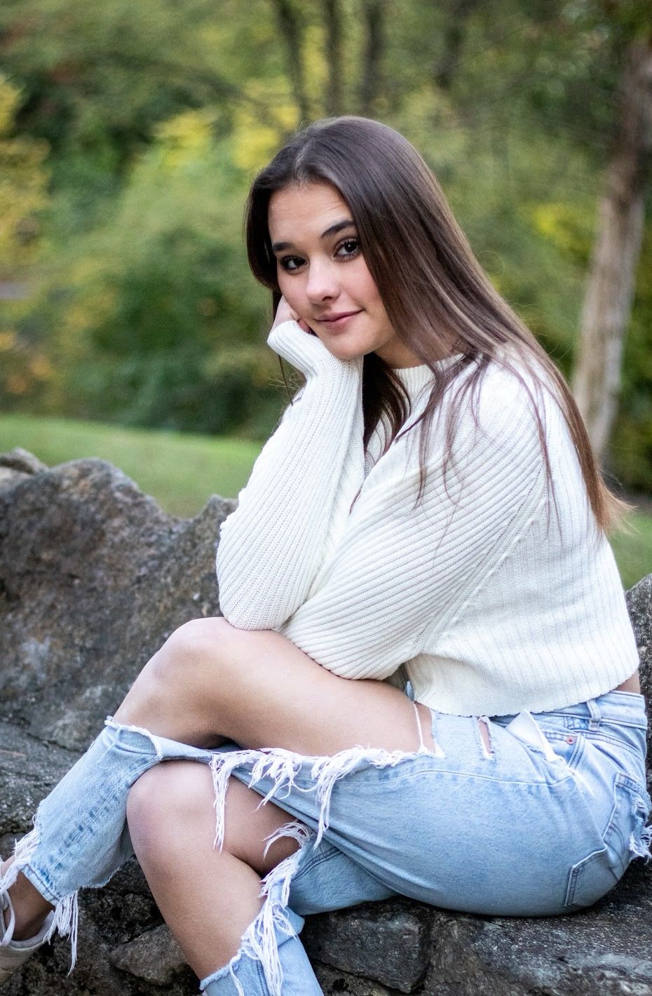 A person in a white sweater and ripped jeans poses casually on rocks outdoors in a natural setting. Senior Session Cincinnati