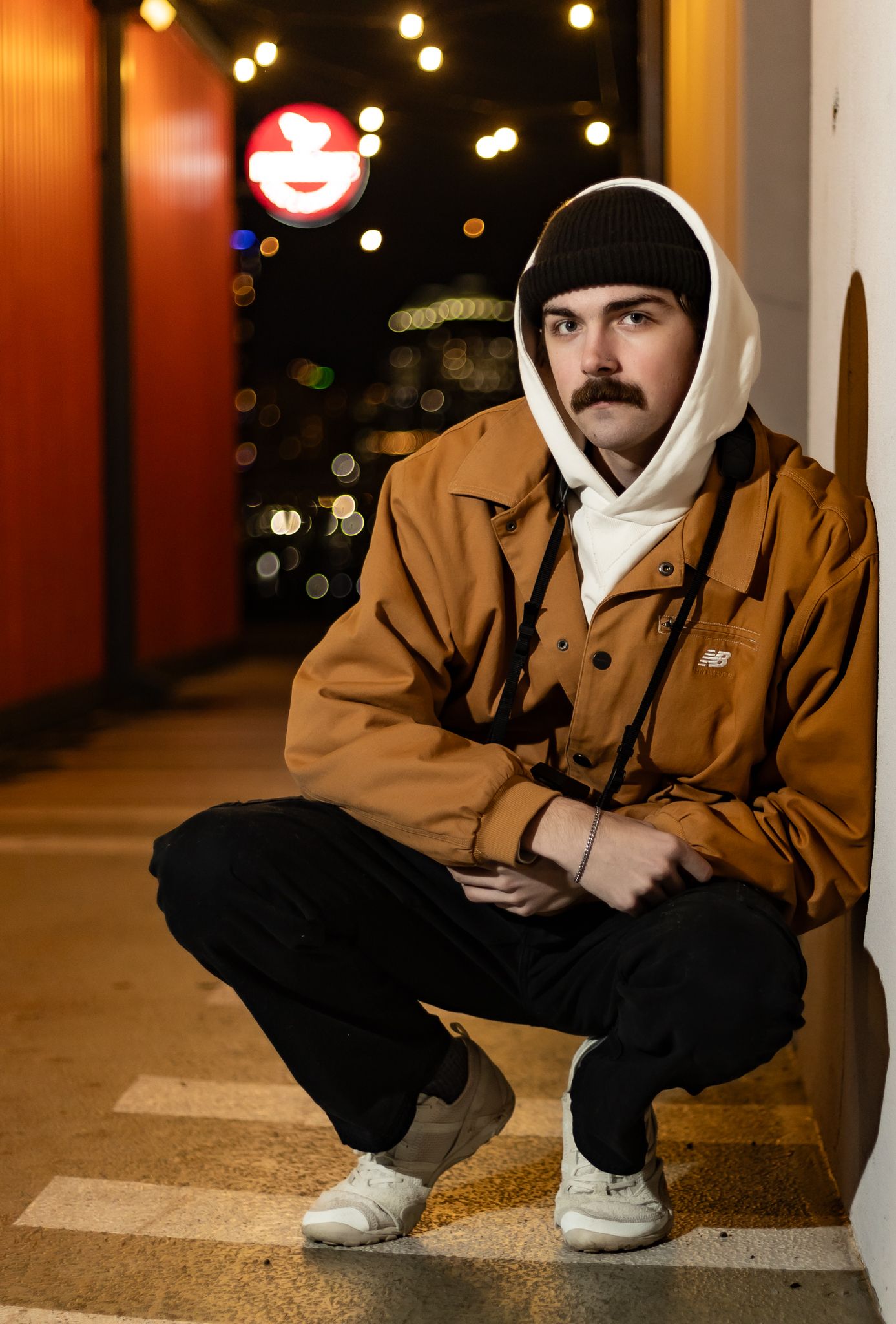 Person in brown jacket and white hoodie crouches against wall with illuminated lights in background. Cincinnati/ northern Kentucky