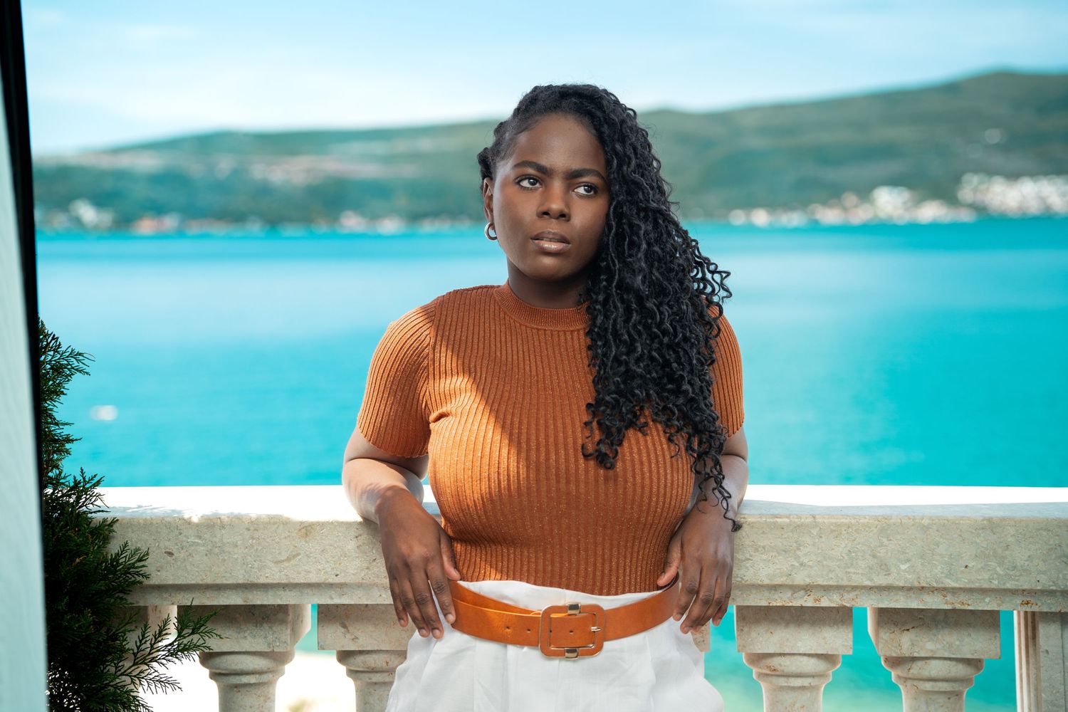 A summertime fashion portrait on a waterfront balcony showcasing a rust-colored top and flowing hair against turquoise waters.