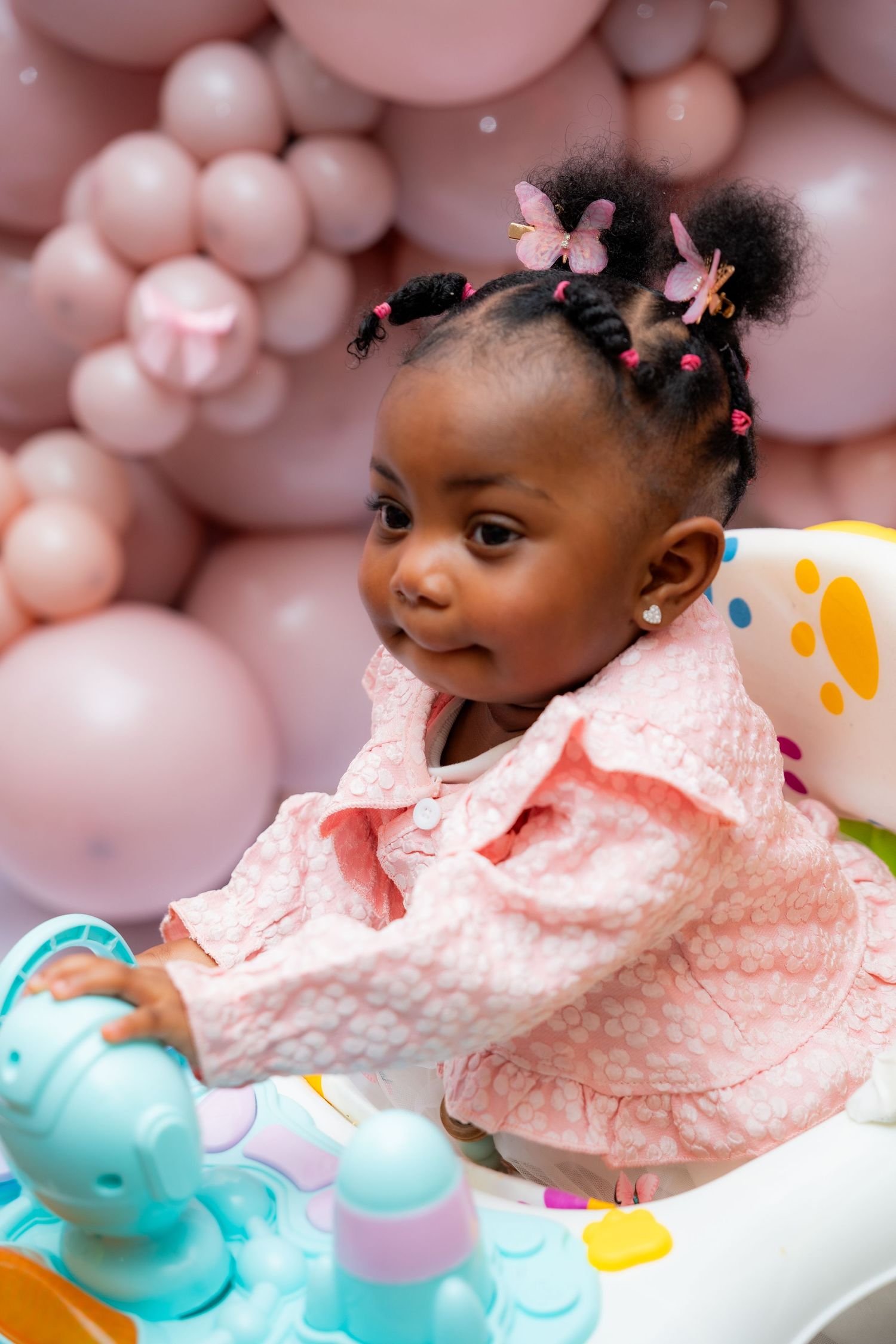 A toddler in a pink dress plays with a tea set against a backdrop of pink balloon decorations.