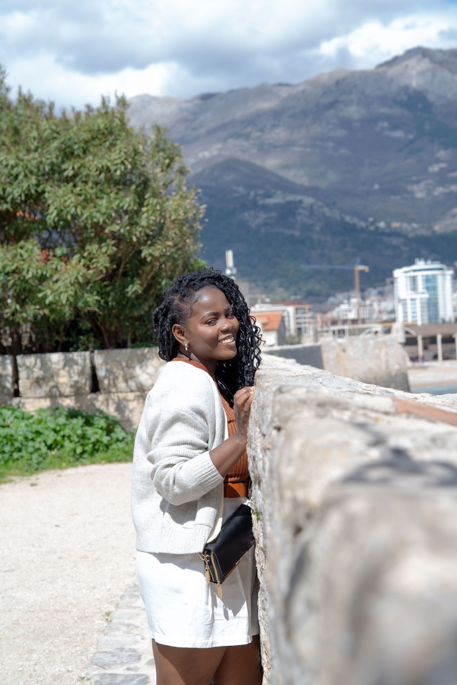 Someone in a white jacket smiling near a stone wall with mountains and city buildings visible in the background.