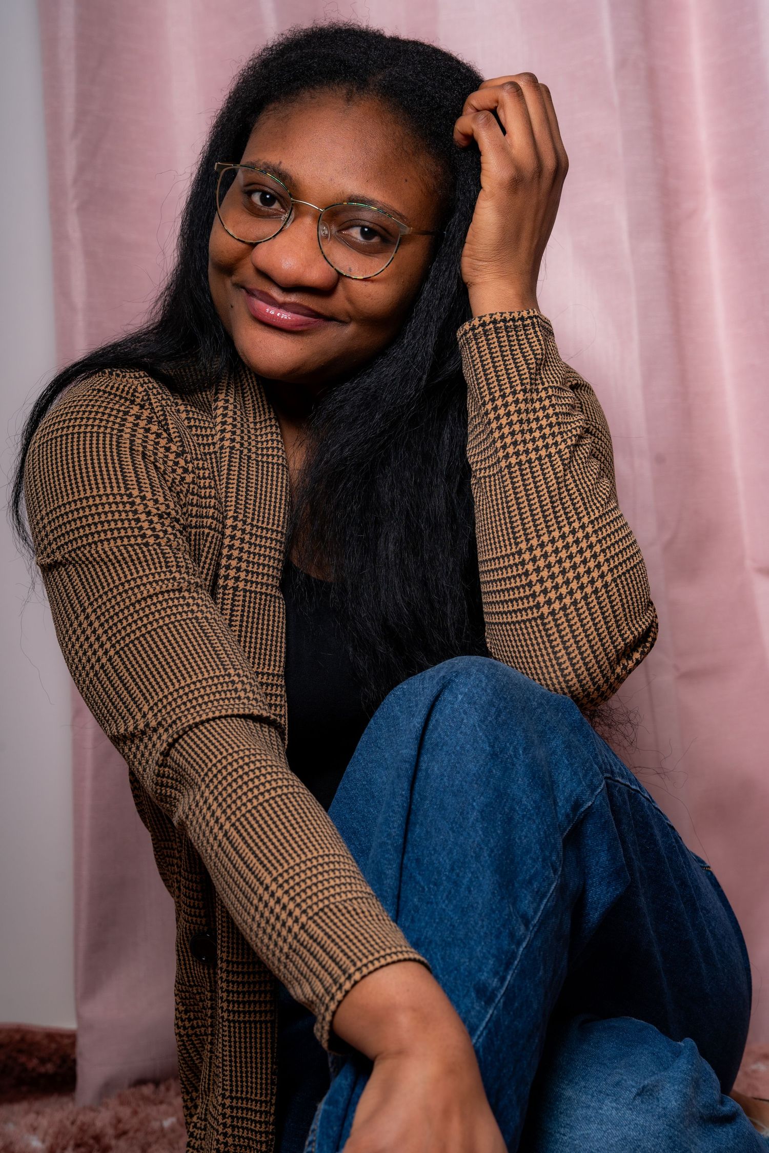 A casual portrait showing a relaxed pose in glasses and plaid jacket against a pink background.