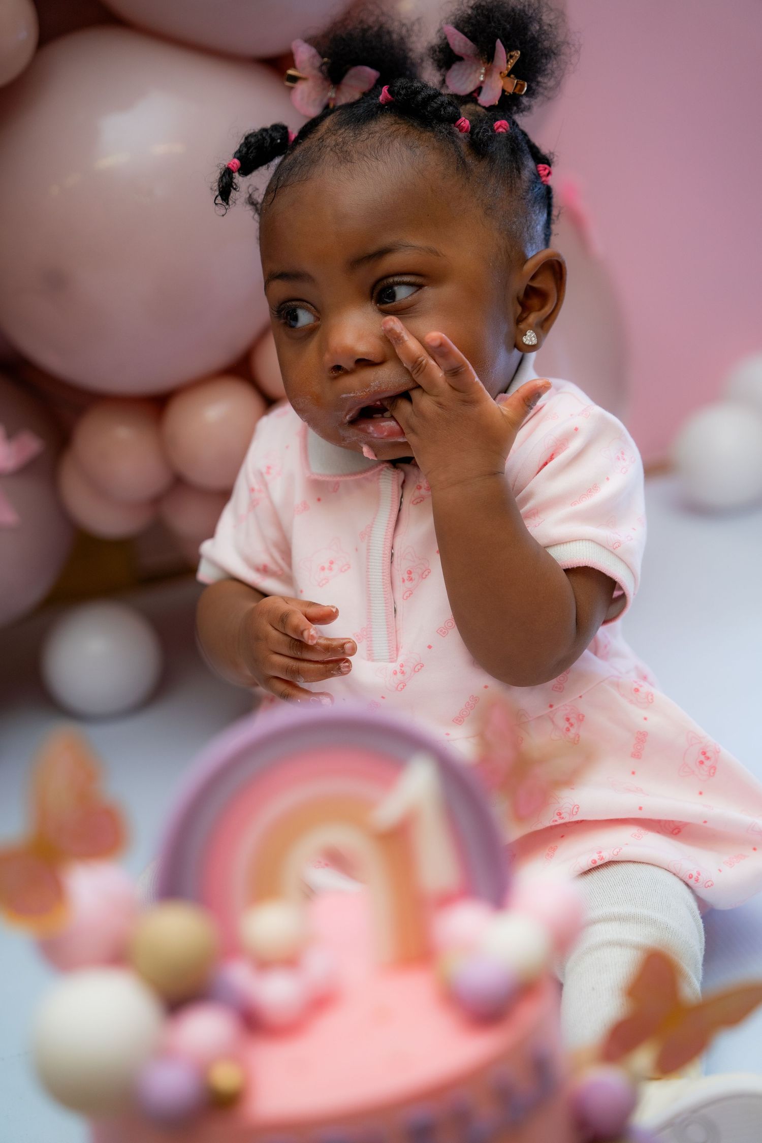 Young child in pink outfit giggles with hand on cheek against pink balloon backdrop at festive celebration.