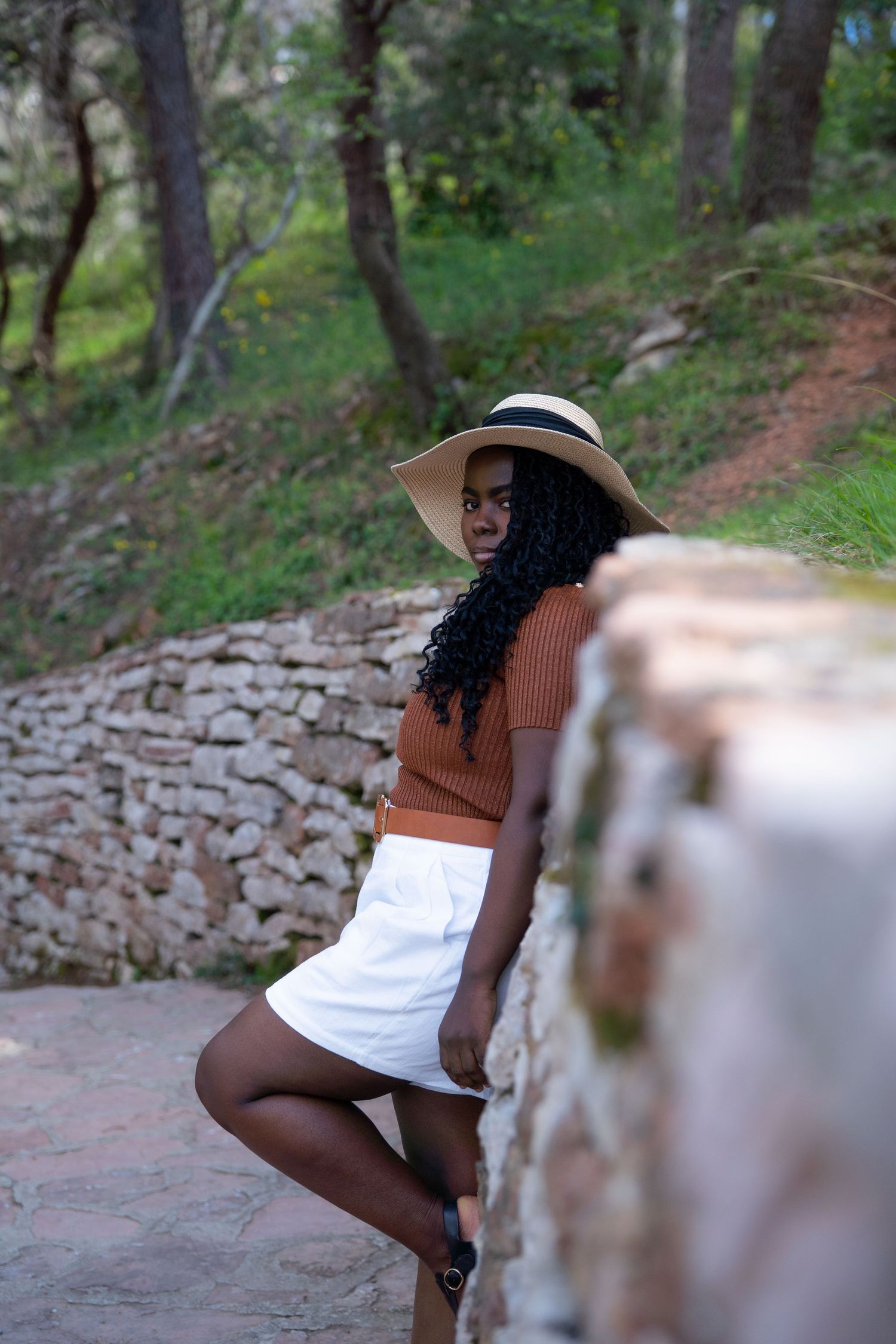 Person wearing a sun hat, orange sweater and white skirt poses on stone steps in a wooded outdoor setting.