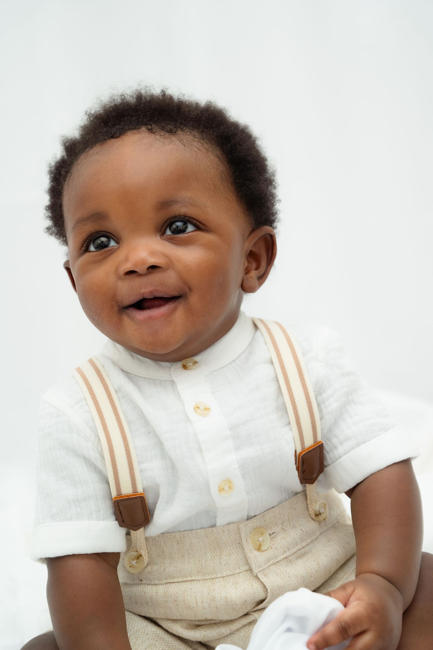 A happy baby wearing a white button-down shirt and tan overalls with suspenders against a light background.