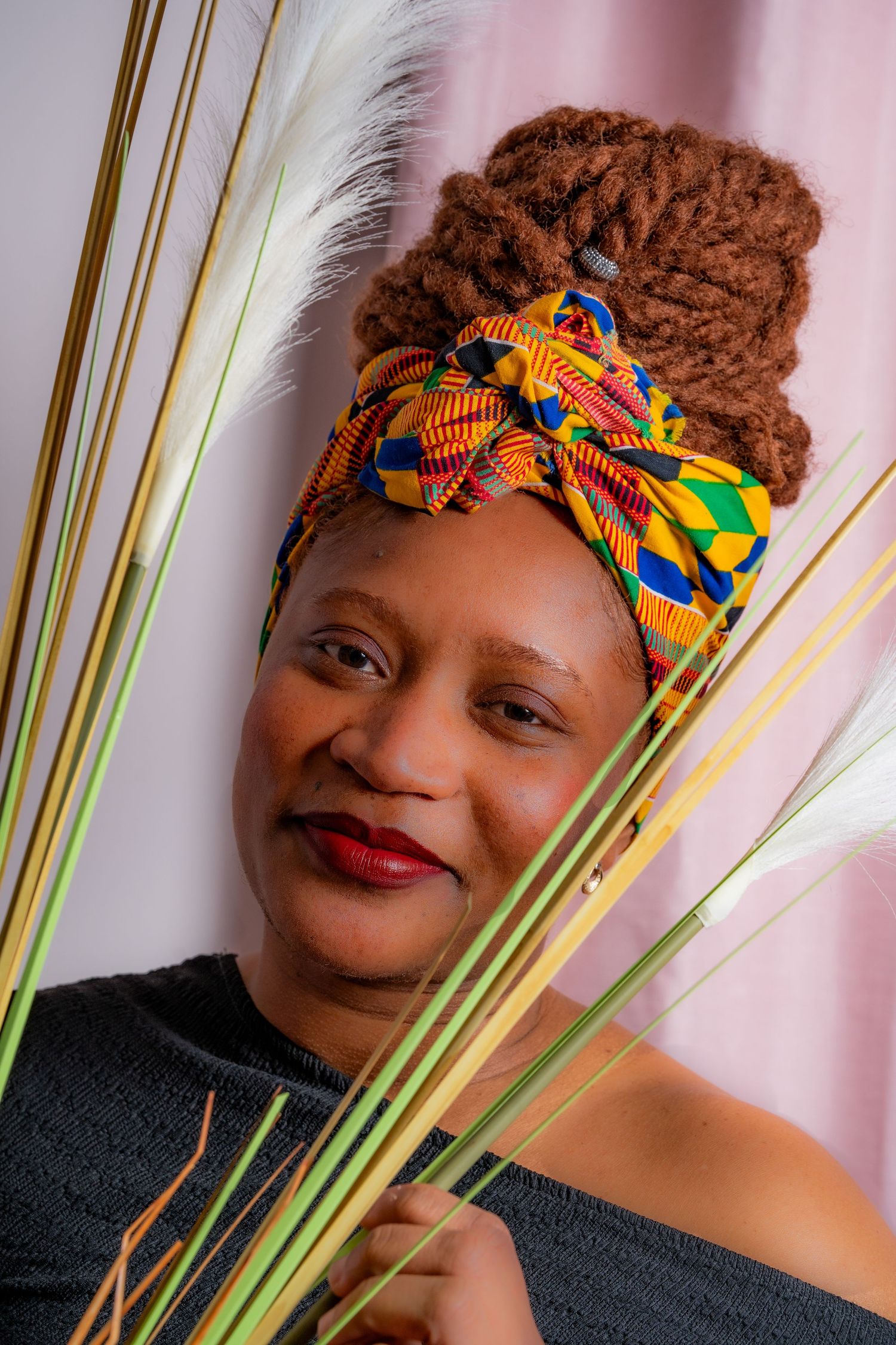 A colorful traditional African headwrap with kente print adorns someone holding green palm fronds against a pink background.