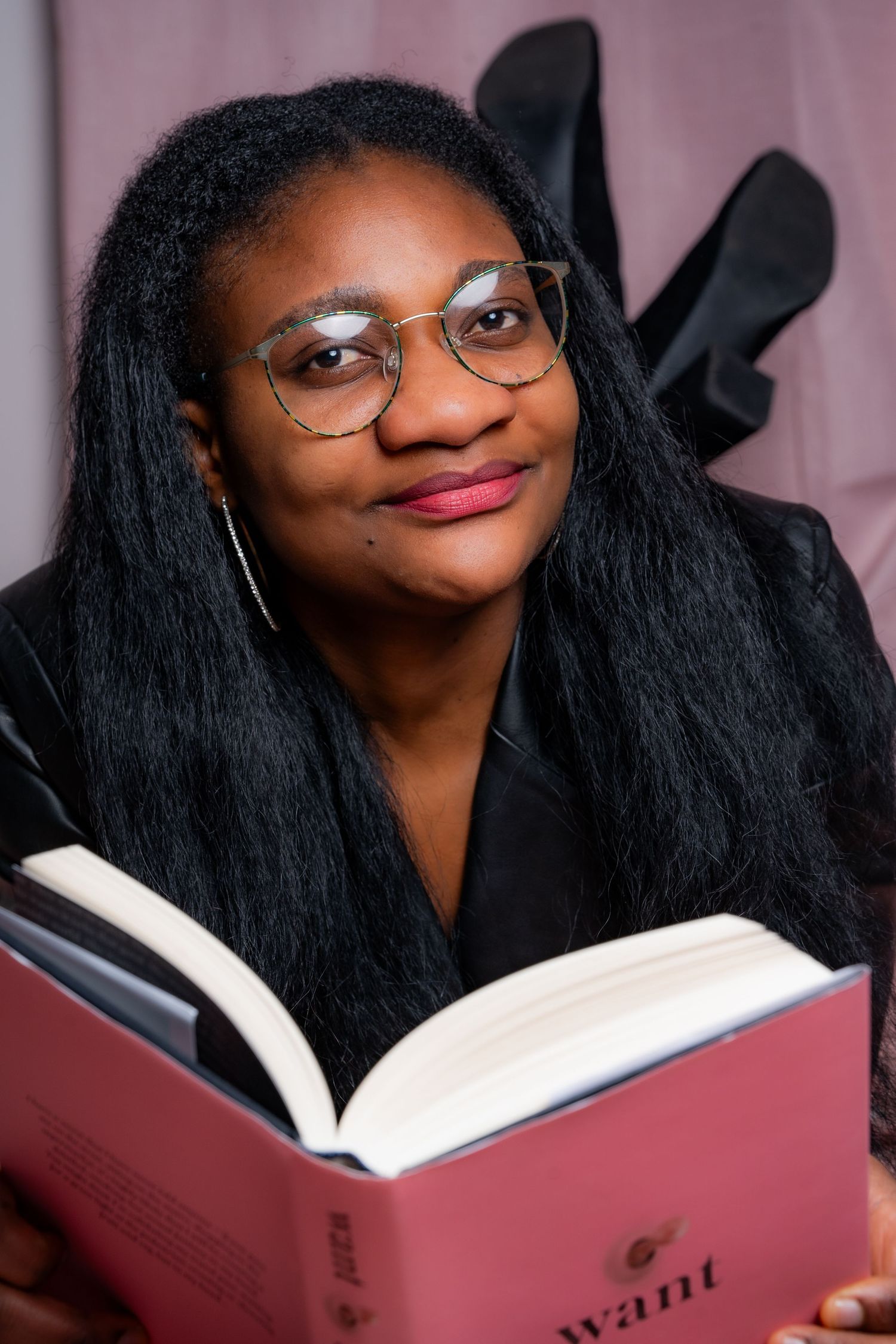 Reader wearing glasses holds an open red book while smiling warmly in front of a dark background.