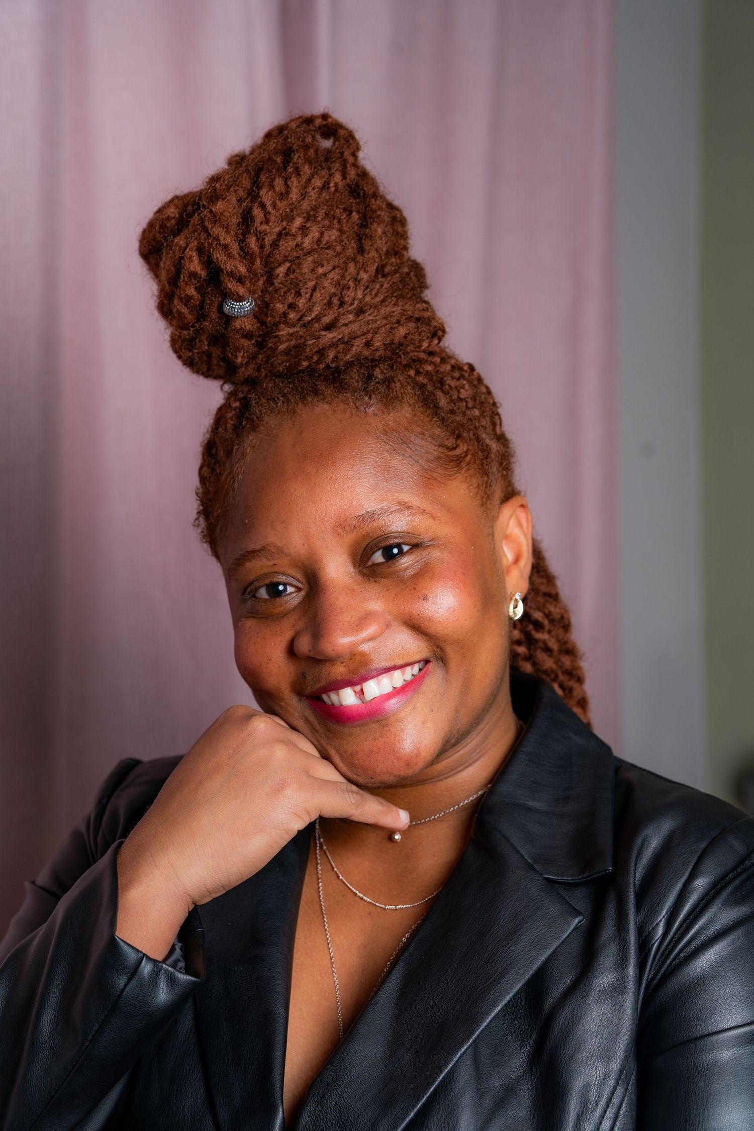 Professional portrait photograph showing bright smile and brown braided updo hairstyle against a pink background.