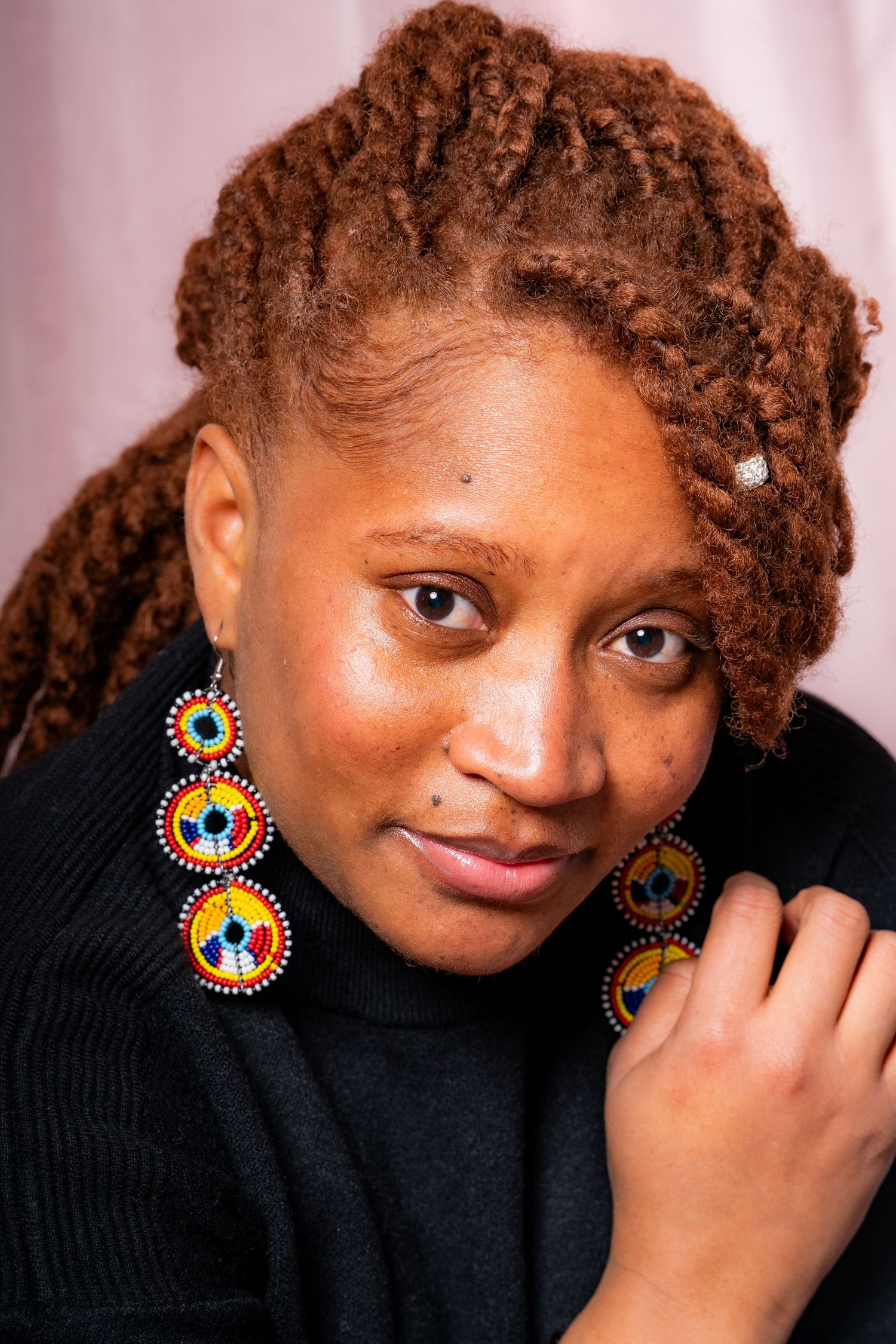 Portrait featuring red-brown dreadlocks and colorful beaded earrings against a soft pink background.