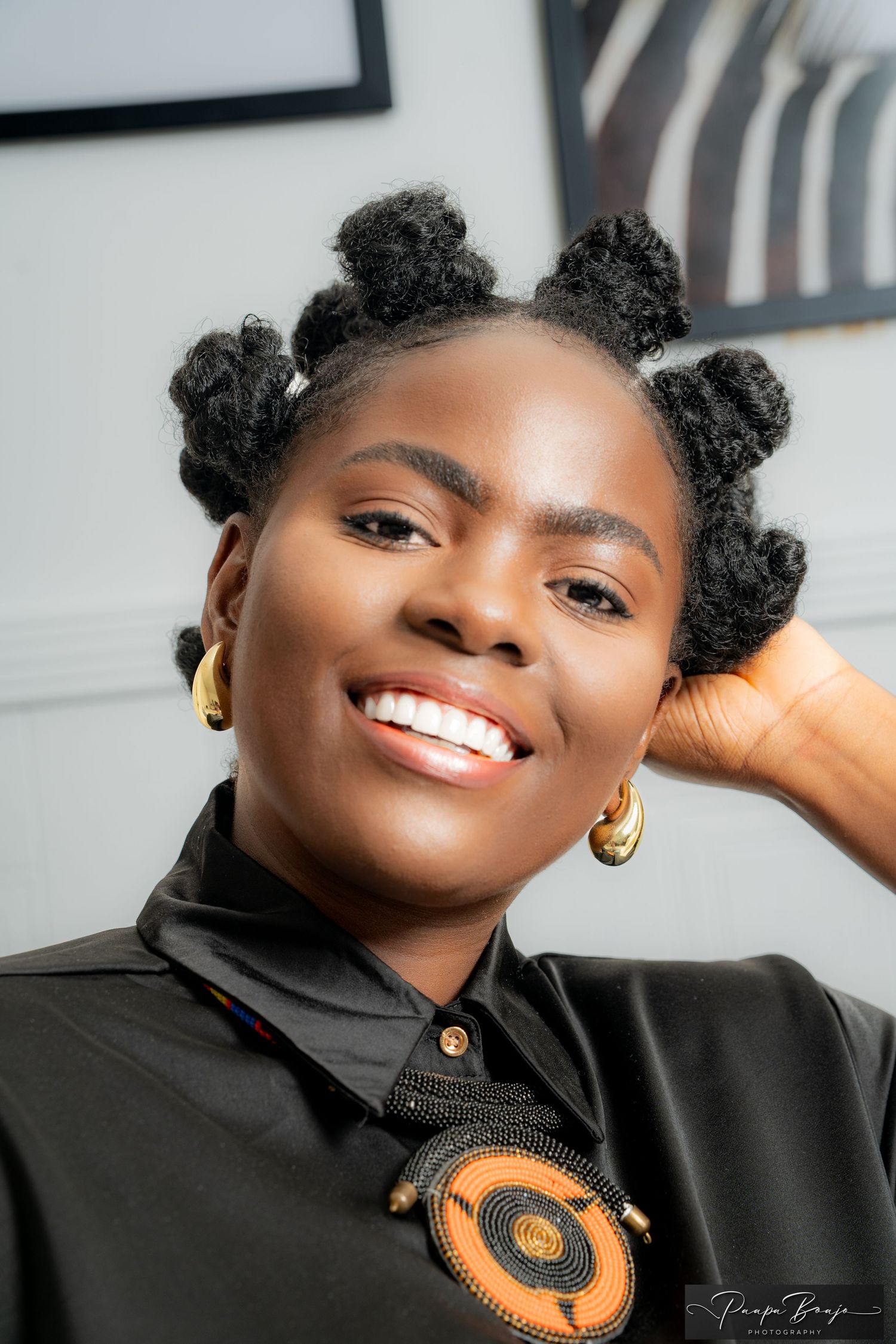 A smiling individual with natural hair styled in bantu knots wearing a black shirt and gold hoop earrings.